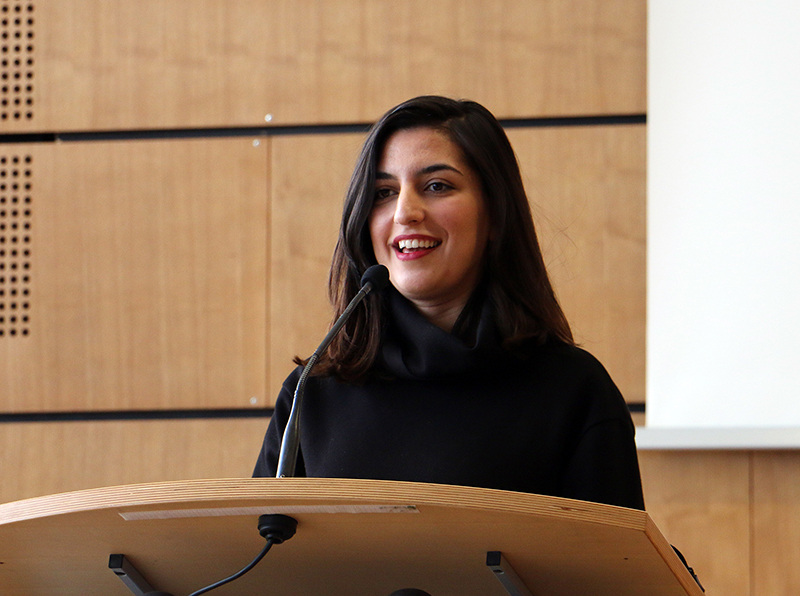 Eine Person mit langen dunklen Haaren lächelt, während sie an einem Podium der Hochschule Coburg spricht. Sie trägt ein schwarzes Outfit und steht vor einem hölzernen Hintergrund mit einem Mikrofon vor sich.