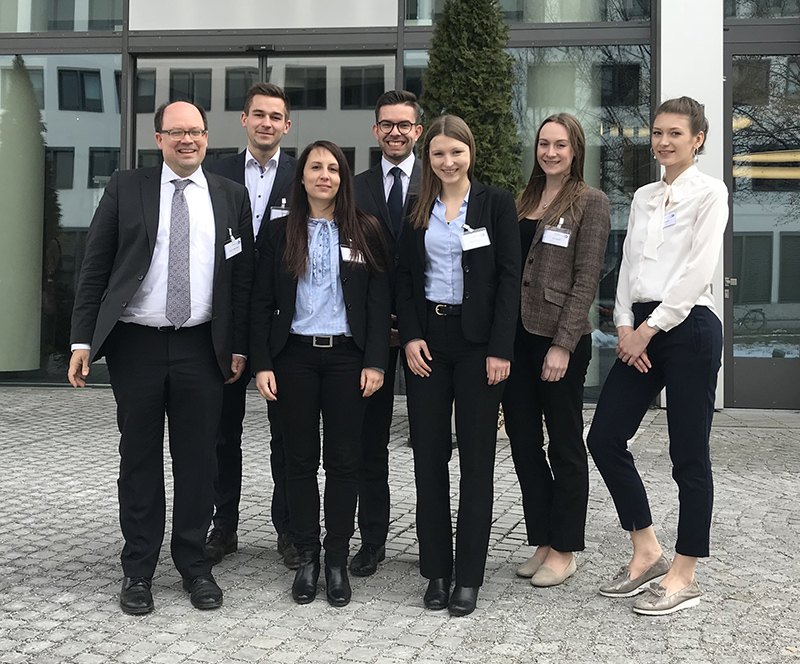 Eine Gruppe von sieben professionell gekleideten Personen steht vor einem Gebäude der Hochschule Coburg und lächelt in die Kamera. Sie tragen Business-Kleidung, darunter Anzüge und Blazer. Im Hintergrund sind Fenster und eine gepflasterte Fläche zu sehen.