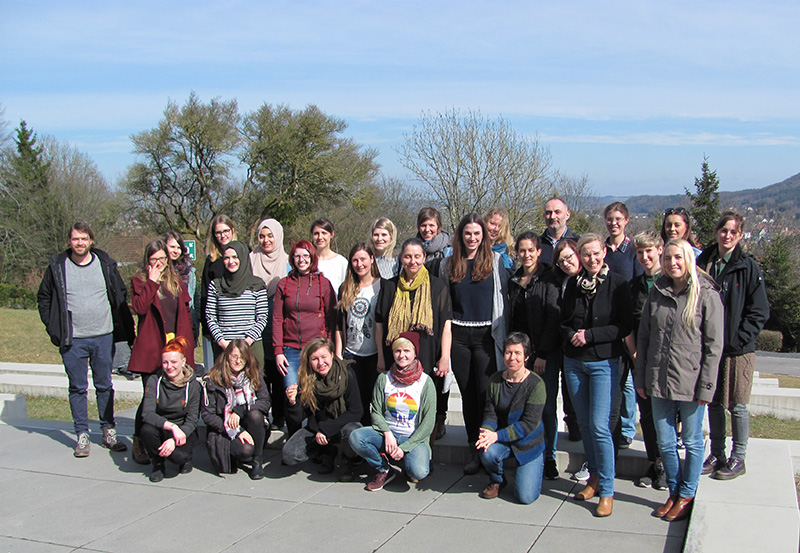 Eine bunt gemischte Gruppe von 27 Personen, die mit der Hochschule Coburg verbunden sind, posiert an einem sonnigen Tag im Freien. Sie stehen und hocken auf einer gepflasterten Fläche mit Bäumen und Hügeln im Hintergrund, lächeln und sind leger gekleidet.