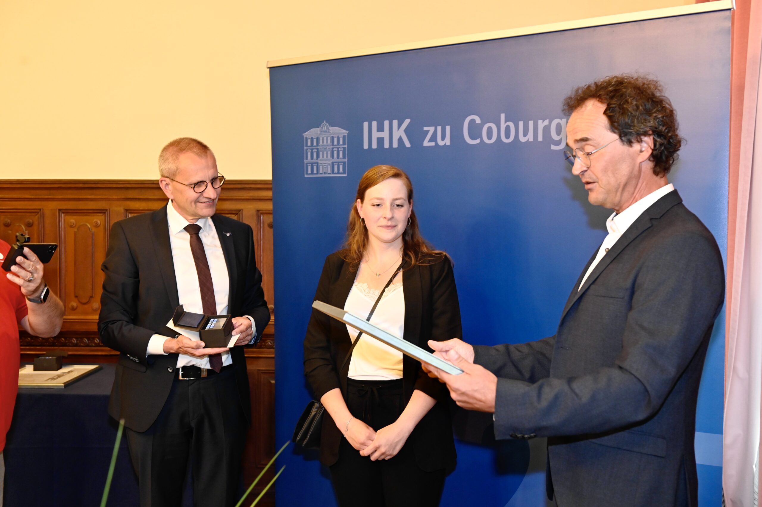 Drei Personen stehen vor einem blauen Banner mit der Aufschrift „IHK zu Coburg“. Ein Mann rechts liest aus einem Dokument vor, während ein Mann und eine Frau links, möglicherweise Angehörige der Hochschule Coburg, aufmerksam zuhören. Der formelle Rahmen lässt auf eine Preisverleihung oder eine Zertifikatsverleihung schließen.