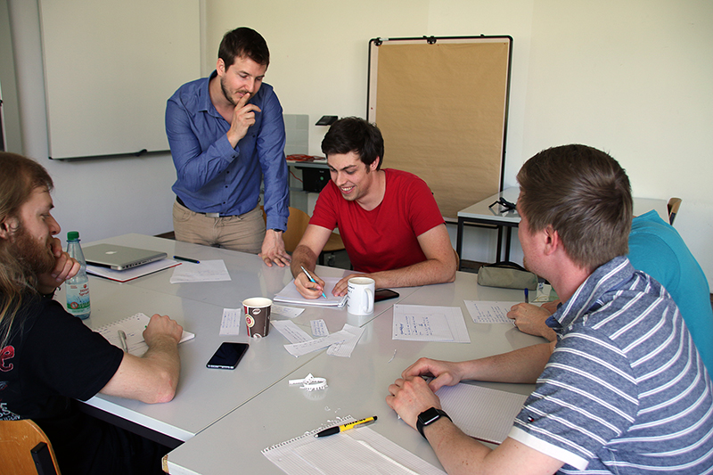 Eine Gruppe von fünf Personen sitzt um einen Tisch in einem Besprechungsraum der Hochschule Coburg. Eine Person steht und unterhält sich, die anderen sitzen, machen sich Notizen und diskutieren. Papiere, Notizbücher und Getränke sind über den Tisch verteilt.