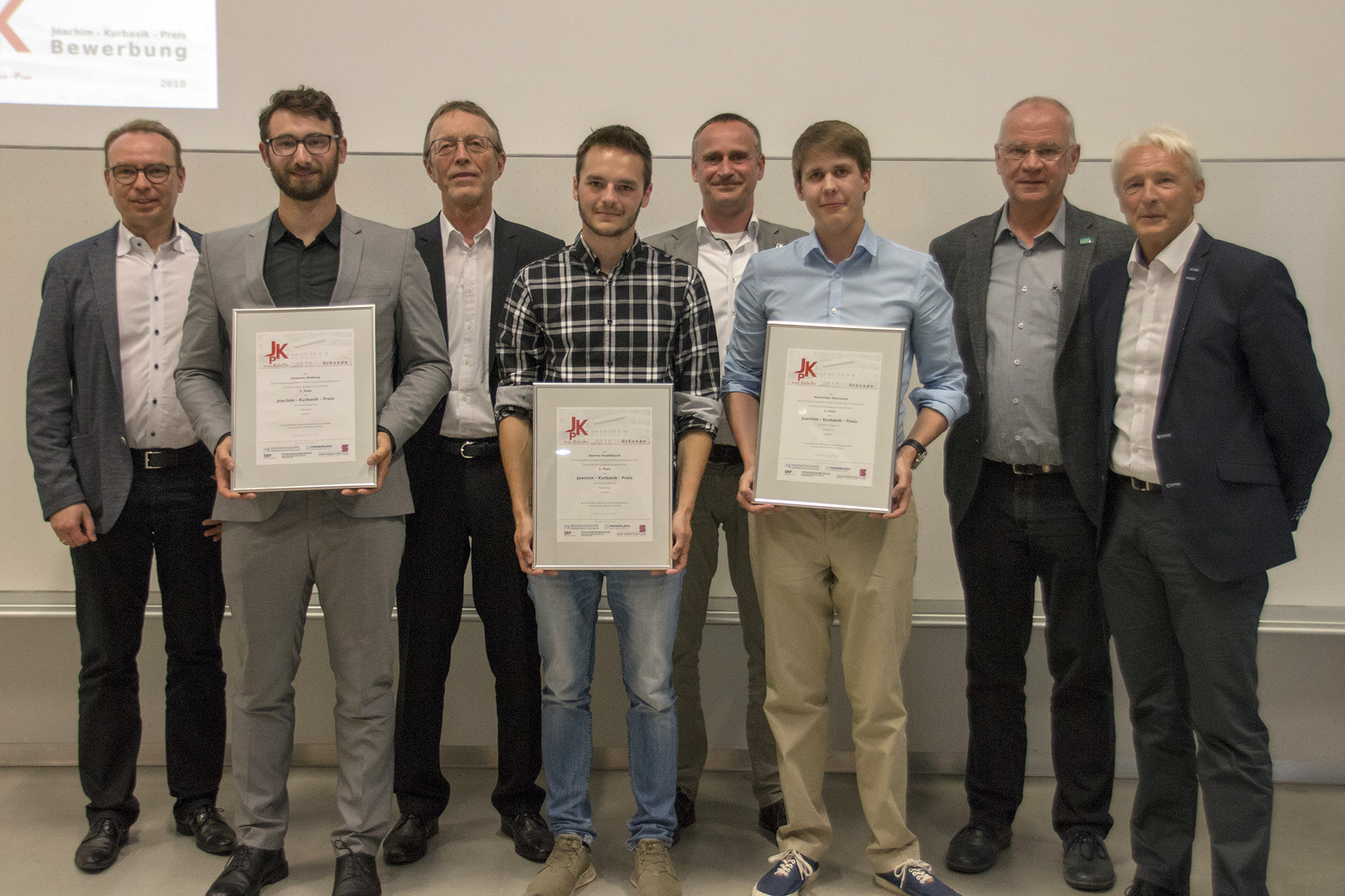 Eine Gruppe von acht Männern steht in einem Gebäude der Hochschule Coburg. Drei von ihnen halten stolz gerahmte Zertifikate in den Händen. Sie sind elegant-leger und formell gekleidet und lächeln vor einem Hintergrund mit einer weißen Tafel und einem Präsentationsbildschirm in die Kamera.