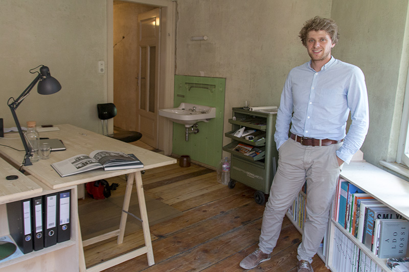 Ein Mann in einem blauen Hemd steht in einem kleinen, minimalistischen Büro mit Holzboden an der Hochschule Coburg. Der Raum verfügt über einen Schreibtisch mit einem aufgeschlagenen Buch, eine Lampe und Regale voller Bücher. Im Hintergrund vervollständigen ein Waschbecken und eine Tür die ruhige akademische Atmosphäre.