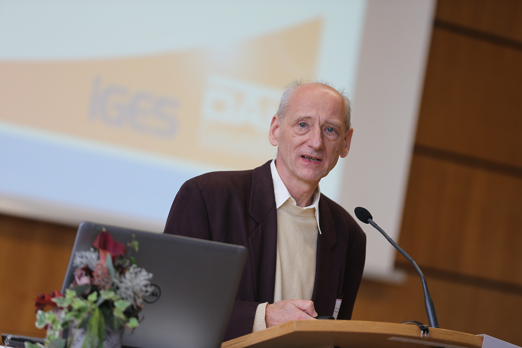 Ein älterer Herr im Anzug spricht an einem Podium mit Mikrofon und vertritt die Hochschule Coburg. Hinter ihm zeigt ein Präsentationsbildschirm undeutliche Logos. Im Vordergrund stehen ein Laptop und ein Blumenarrangement, das den Tisch schmückt.