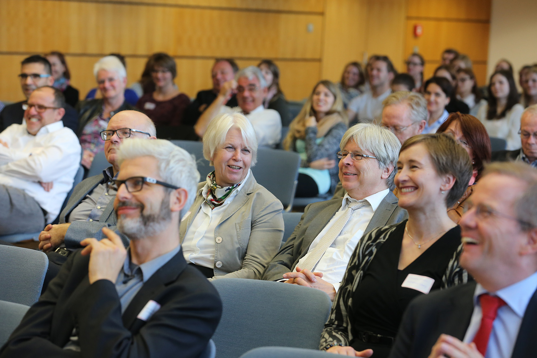 Eine bunt gemischte Gruppe von Menschen sitzt in einem Hörsaal der Hochschule Coburg, hört aufmerksam zu und lächelt. In legerer Business-Kleidung gekleidet, lassen sie eine berufliche oder lehrreiche Veranstaltung vermuten. Das Ambiente ist modern, mit Holzwänden und Stuhlreihen.