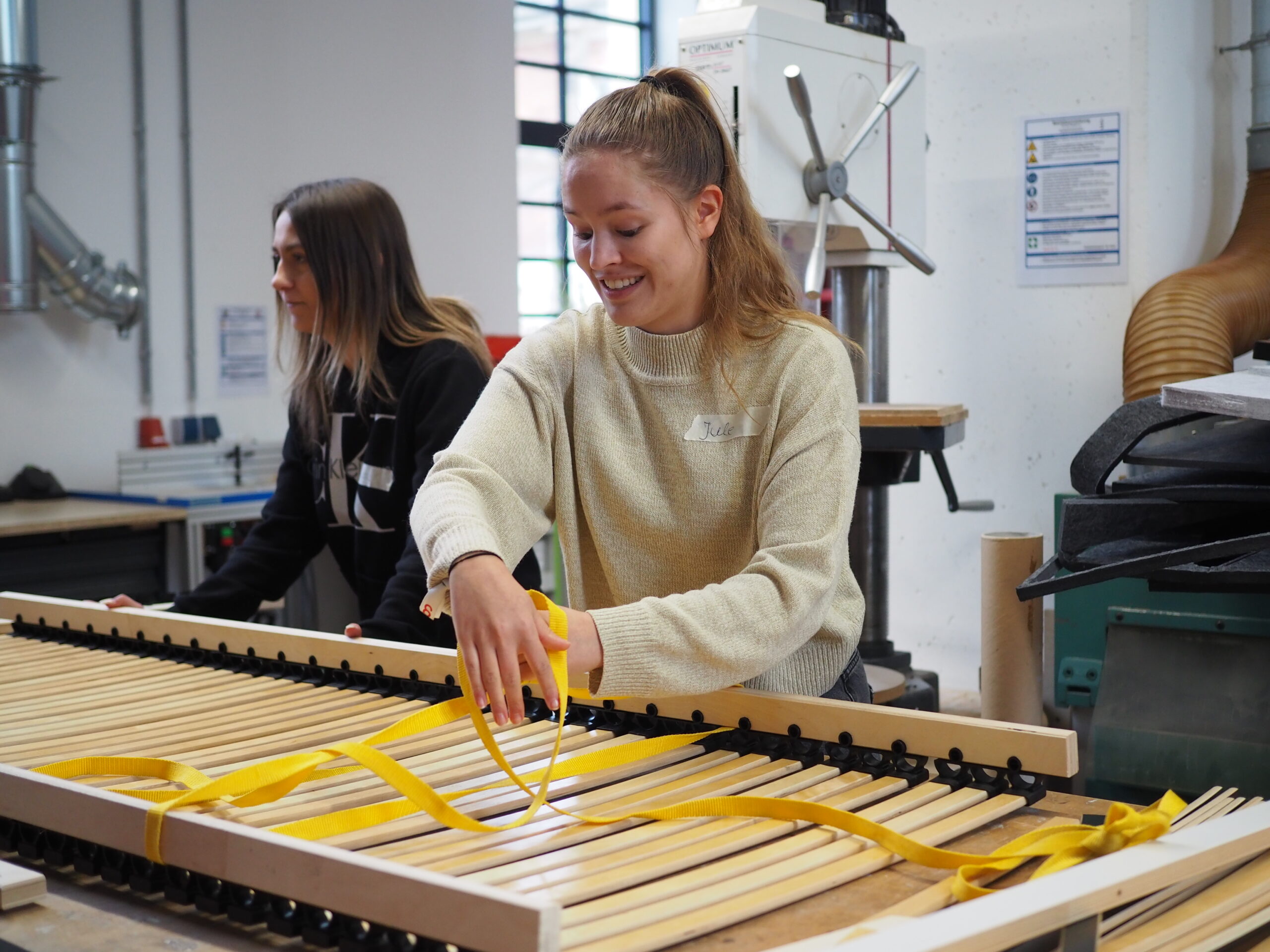 In einer geschäftigen Werkstatt der Hochschule Coburg montieren zwei Personen ein Holzstück mit gelben Bändern. Einer, in einem cremefarbenen Pullover, lächelt selbstbewusst, während er die Bänder handhabt, während der andere inmitten der um sie herum verstreuten Industriegeräte und Werkzeuge Halt bietet.