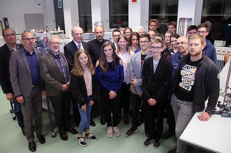 Eine Gruppe von Menschen unterschiedlichen Alters steht in einem gut beleuchteten Raum der Hochschule Coburg. Einige sind leger gekleidet, andere tragen Business-Kleidung. Sie scheinen sich in einem akademischen oder beruflichen Umfeld zu befinden, im Hintergrund sind Tische und Geräte zu sehen.