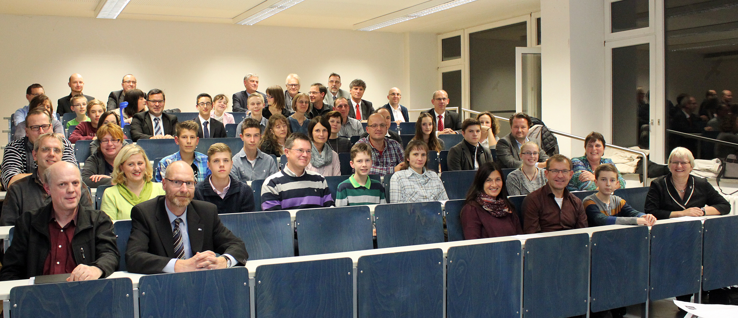 Eine große Gruppe von Menschen, darunter Männer, Frauen und Kinder, sitzt in einem Hörsaal der Hochschule Coburg. Sie sind alle in legerer Business-Kleidung gekleidet. Der Raum ist mit Reihen blauer Schreibtische und weißen Wänden mit großen Fenstern ausgestattet. Jeder blickt in die Kamera und lächelt strahlend.