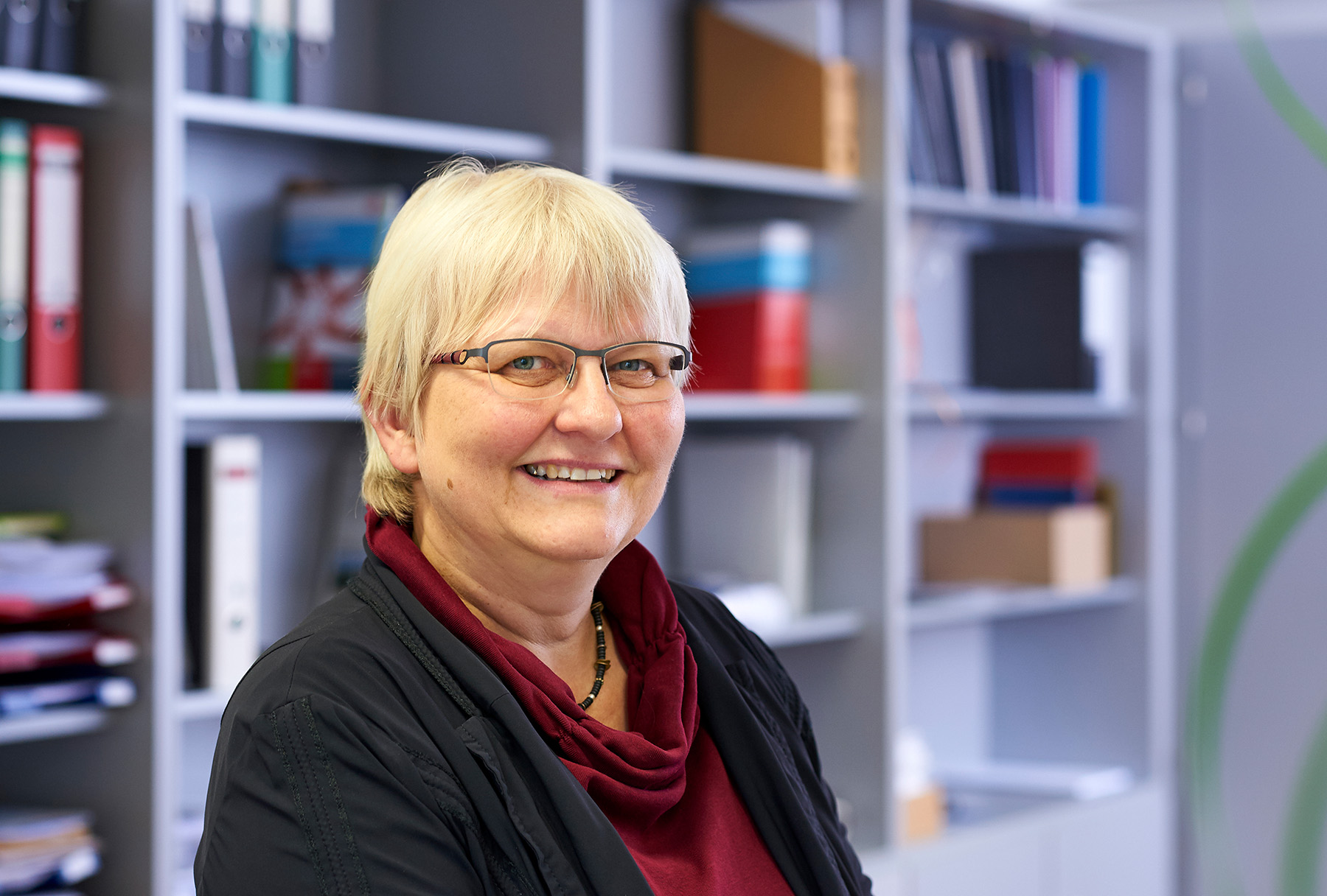 Eine Person mit kurzen blonden Haaren und Brille steht lächelnd vor einem Regal voller Bücher und Ordner in der Hochschule Coburg. Sie trägt eine schwarze Jacke und ein kastanienbraunes Oberteil.