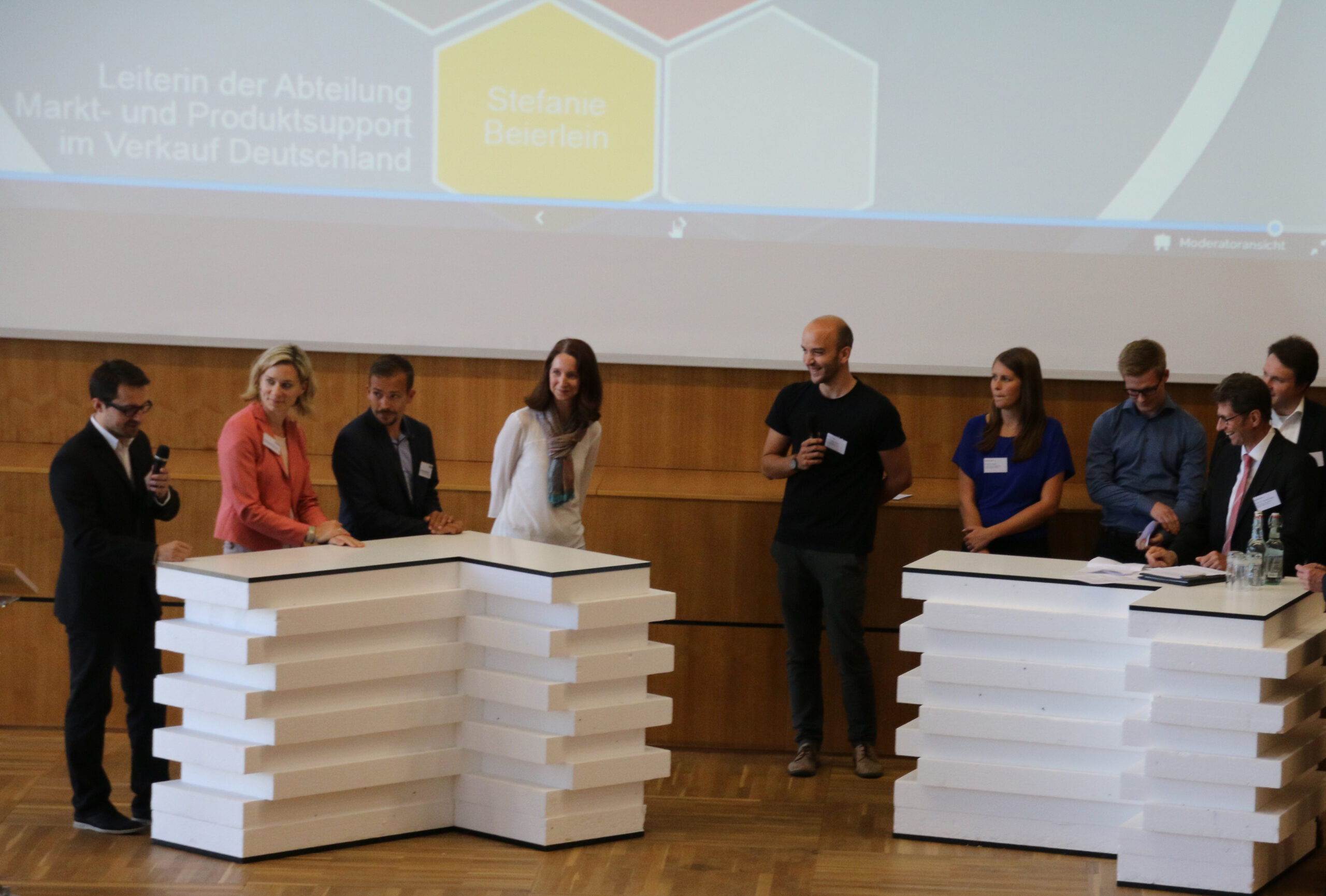 Eine Gruppe von Menschen steht auf einer Bühne mit zwei weißen Podien und nimmt an einer Podiumsdiskussion teil. Im Hintergrund ist eine Präsentationsfolie zu sehen, die darauf hinweist, dass es sich um eine akademische Veranstaltung der Hochschule Coburg handelt. Das Setting erinnert an eine Konferenz oder ein Seminar.