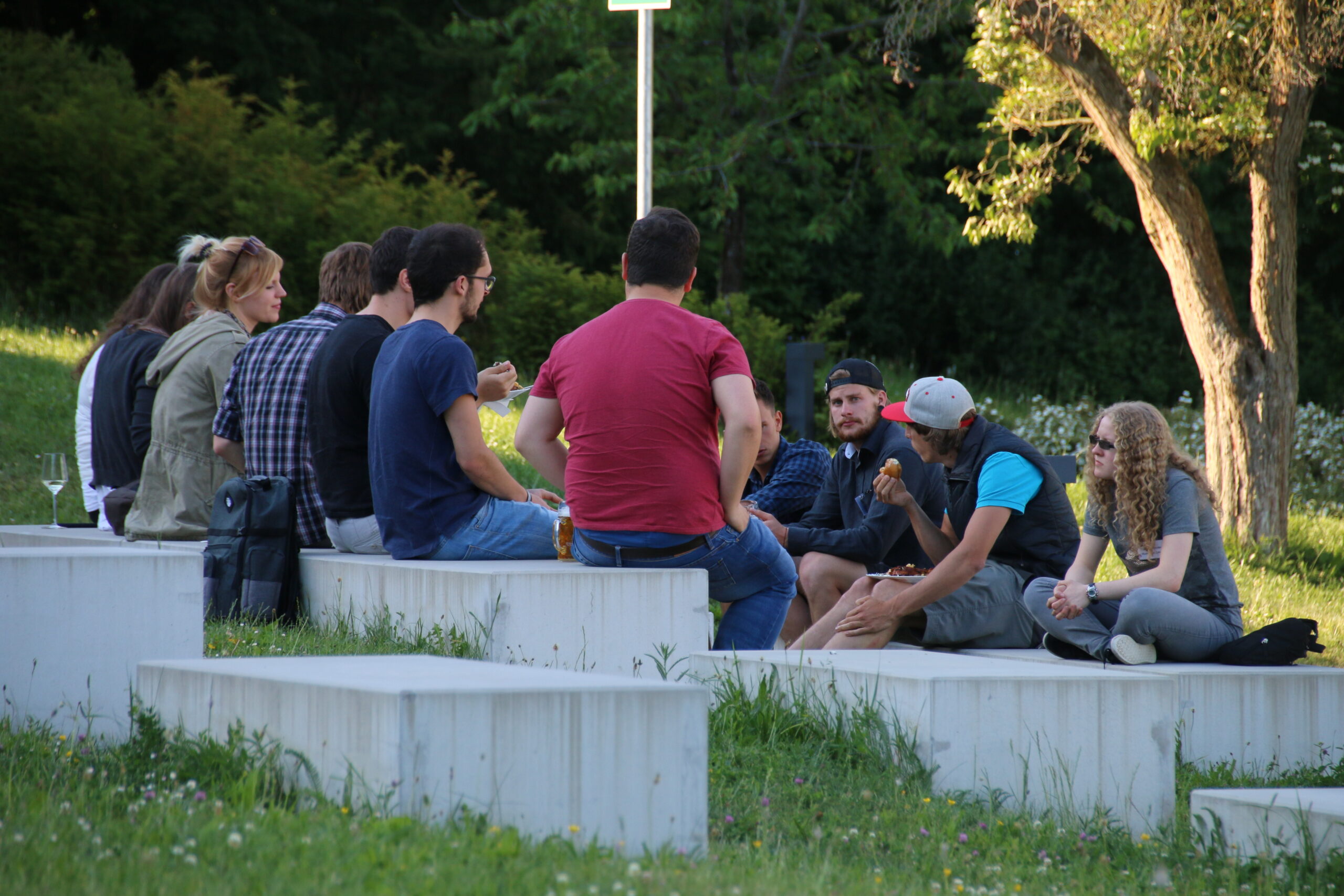 Eine Gruppe von Menschen sitzt draußen auf Betonbänken in einem Park in der Nähe der Hochschule Coburg und unterhält sich angeregt. Der Bereich ist von Gras und Bäumen umgeben, was eine ruhige und natürliche Umgebung schafft. Einige Teilnehmer gestikulieren lebhaft, während sie sprechen.