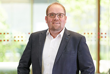 Ein Mann mit Brille steht in einem Gebäude der Hochschule Coburg. Er trägt einen dunklen Blazer über einem weißen Hemd. Vor dem hellen Hintergrund unscharfer Fenster blickt er mit neutralem Gesichtsausdruck in die Kamera.