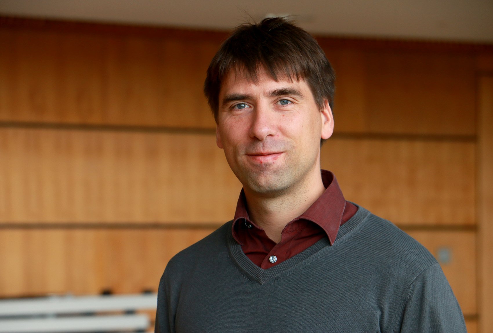 Eine Person mit kurzen braunen Haaren, die einen grauen Pullover über einem kastanienbraunen Hemd trägt, steht vor einer holzgetäfelten Wand in der Hochschule Coburg und lächelt leicht in die Kamera.