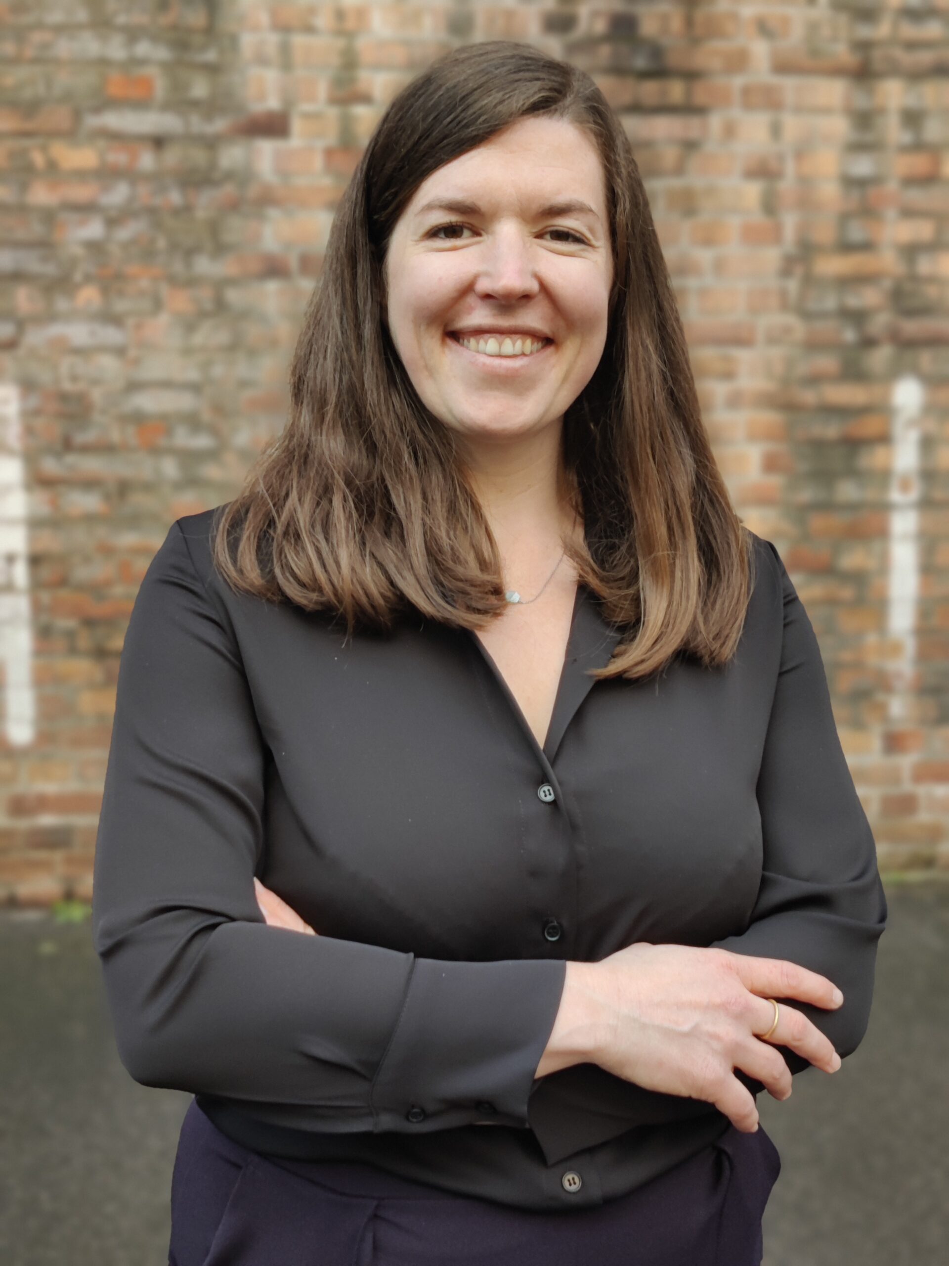 A woman with long brown hair stands smiling with arms crossed, wearing a black blouse. She is in front of a brick wall, exuding confidence and embodying the spirit of Hochschule Coburg.