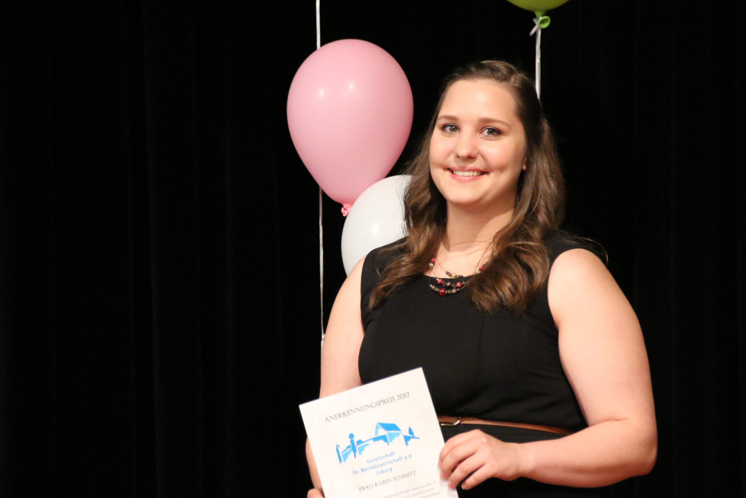 Eine Frau in einem schwarzen Kleid von der Hochschule Coburg lächelt, während sie eine Urkunde in der Hand hält und vor einem schwarzen Hintergrund steht, der mit rosa und weißen Luftballons geschmückt ist.