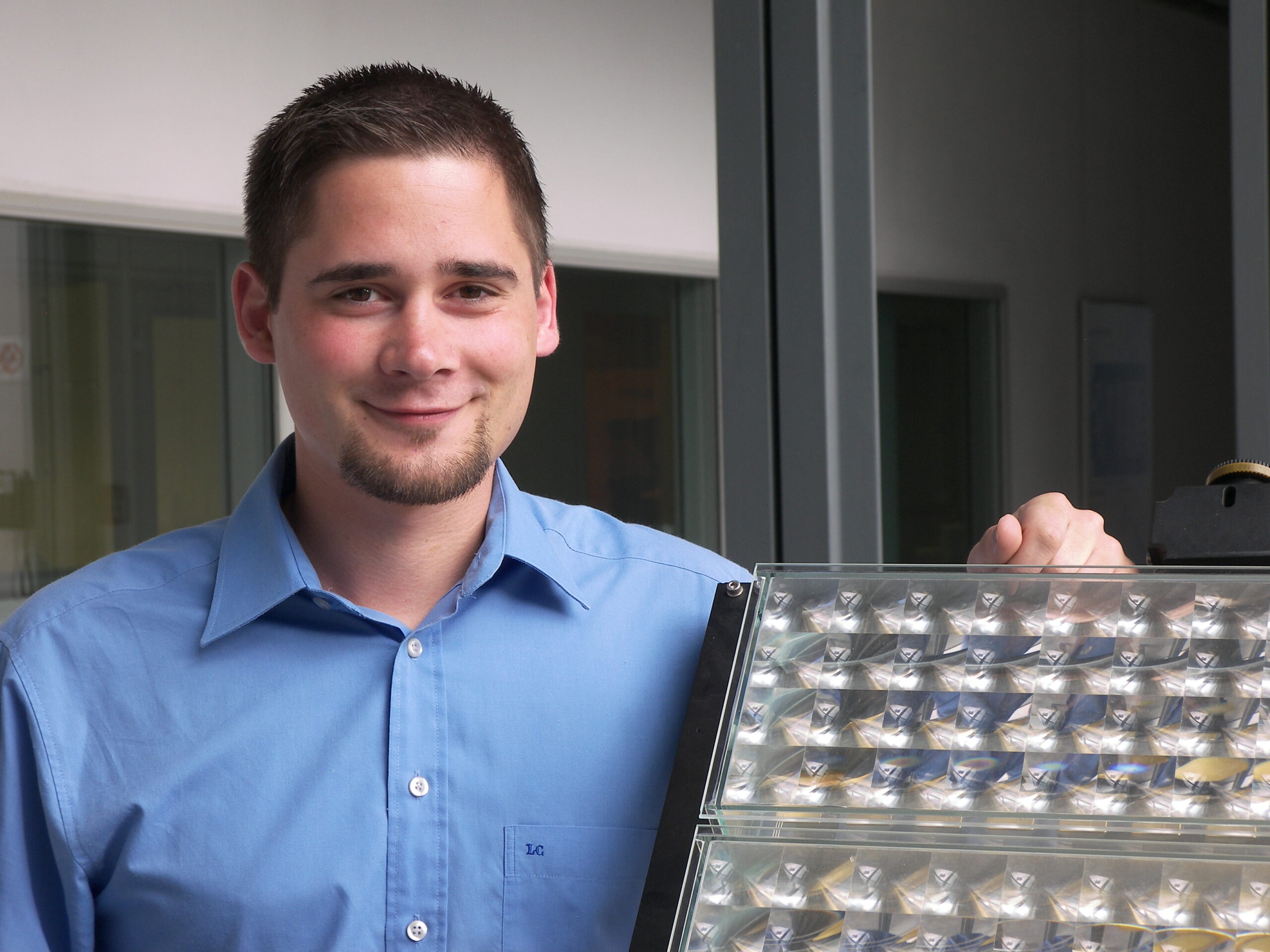 Ein Mann in einem blauen Hemd, möglicherweise ein Absolvent der Hochschule Coburg, lächelt neben einer großen Tafel mit mehreren reflektierenden Quadraten. Er hat kurze Haare und einen gestutzten Bart. Im Hintergrund sind Bürofenster und eine graue Wand zu sehen, was auf die moderne Architektur der Hochschule hindeutet.