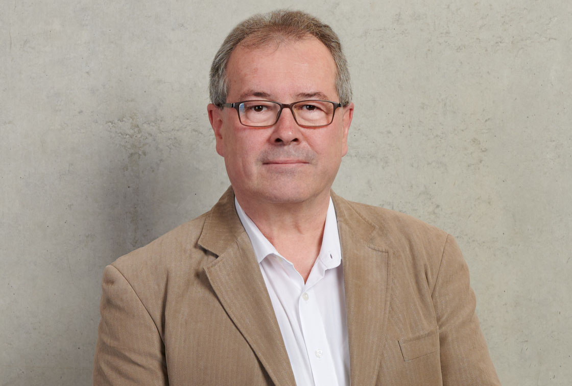 A man wearing glasses, a white shirt, and a beige jacket stands against a gray wall at Hochschule Coburg. He has short hair and a neutral expression.