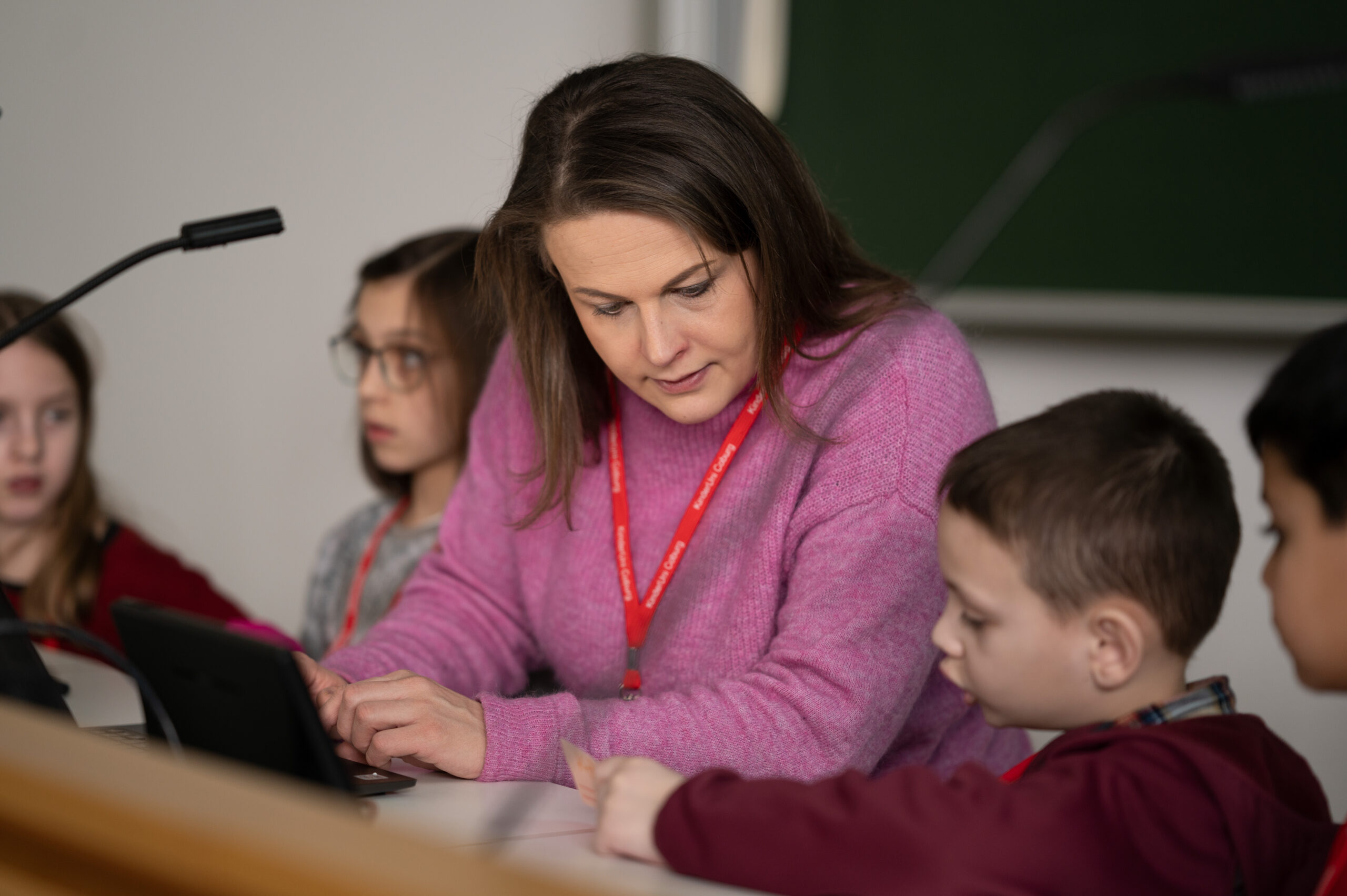 Eine Frau in einem rosa Pullover, an dem ein Schlüsselband der Hochschule Coburg hängt, hilft einem kleinen Jungen an einem Schreibtisch bei den Hausaufgaben. Andere Kinder sitzen in der Nähe und sind in ihre Aufgaben vertieft. Als Hintergrund dient eine große grüne Tafel.