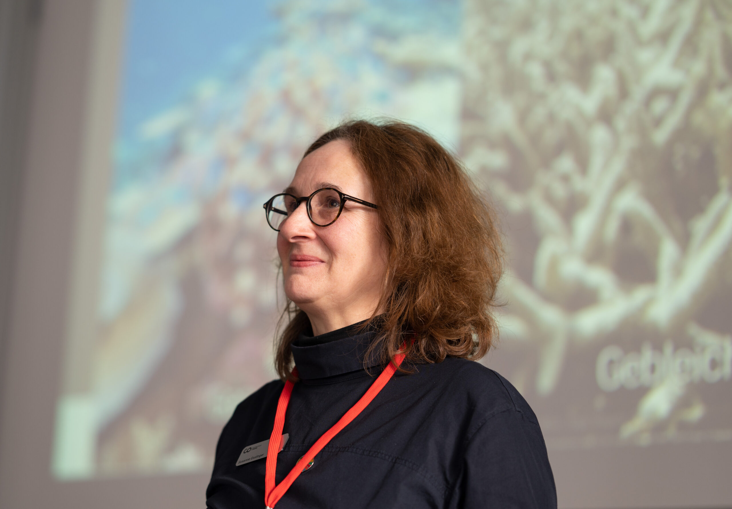 Eine Person mit schulterlangem Haar und Brille, einem dunklen Oberteil und einem roten Schlüsselband steht vor einem unscharfen Hintergrund mit einem abstrakten Bild, möglicherweise an der Hochschule Coburg.