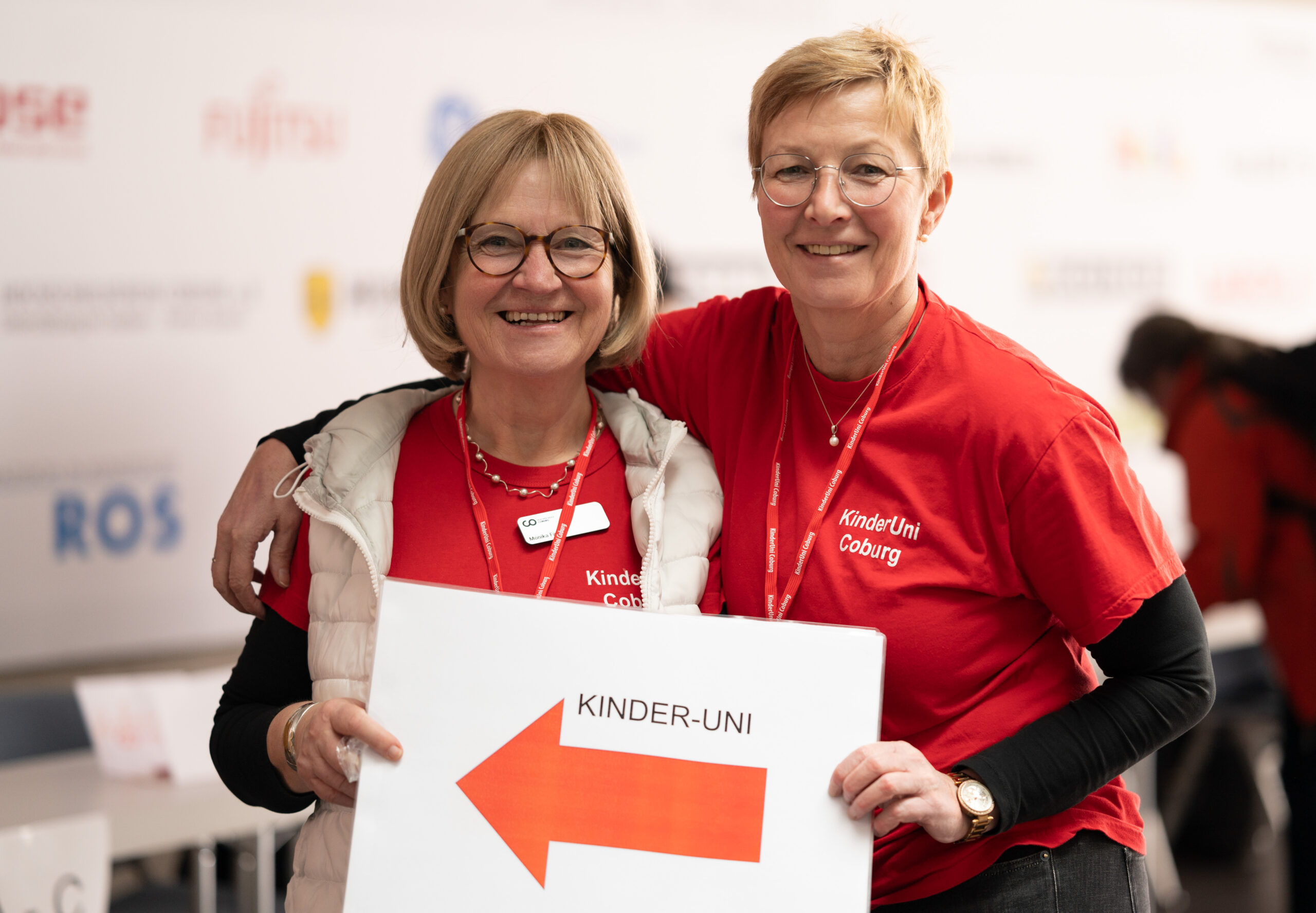 Zwei lächelnde Frauen in roten „Kinder-Uni Coburg“-Shirts stehen stolz mit einem Schild da, auf dem ein großer orangefarbener Pfeil nach links zeigt. Sie posieren vor einem Hintergrund, der mit verschiedenen Logos geschmückt ist, die die Hochschule Coburg repräsentieren.