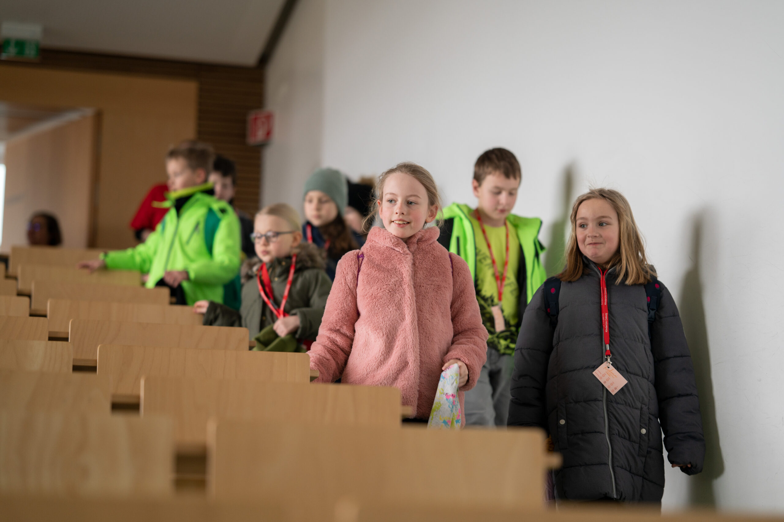 Eine Gruppe Kinder in Wintermänteln, möglicherweise zukünftige Stipendiaten der Hochschule Coburg, geht in einem Hörsaal die Treppe hinunter. Sie tragen bunte Jacken und rote Umhängebänder, tragen Taschen und wirken fröhlich und engagiert. Der Raum hat Holzsitze und eine weiße Wand.