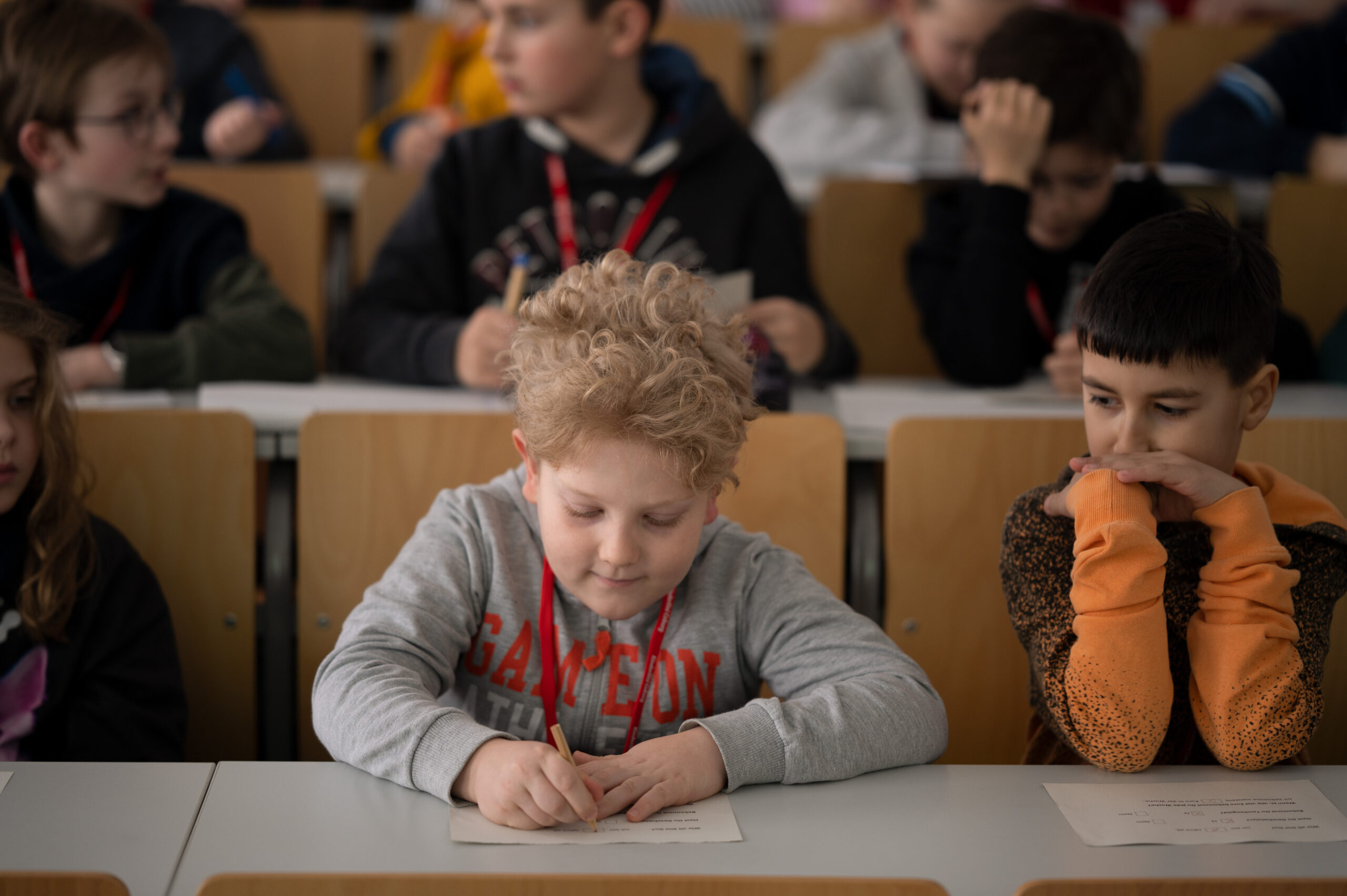 In einer lebhaften Unterrichtsszene sitzen junge Schüler an ihren Schreibtischen und sind ins Lernen vertieft. Ein Kind mit lockigem Haar im Vordergrund konzentriert sich aufs Schreiben und verkörpert damit den akademischen Geist der Hochschule Coburg. Andere Kinder blicken sich um oder schauen auf ihre Arbeit. Alle tragen Freizeitkleidung und Schlüsselbänder.