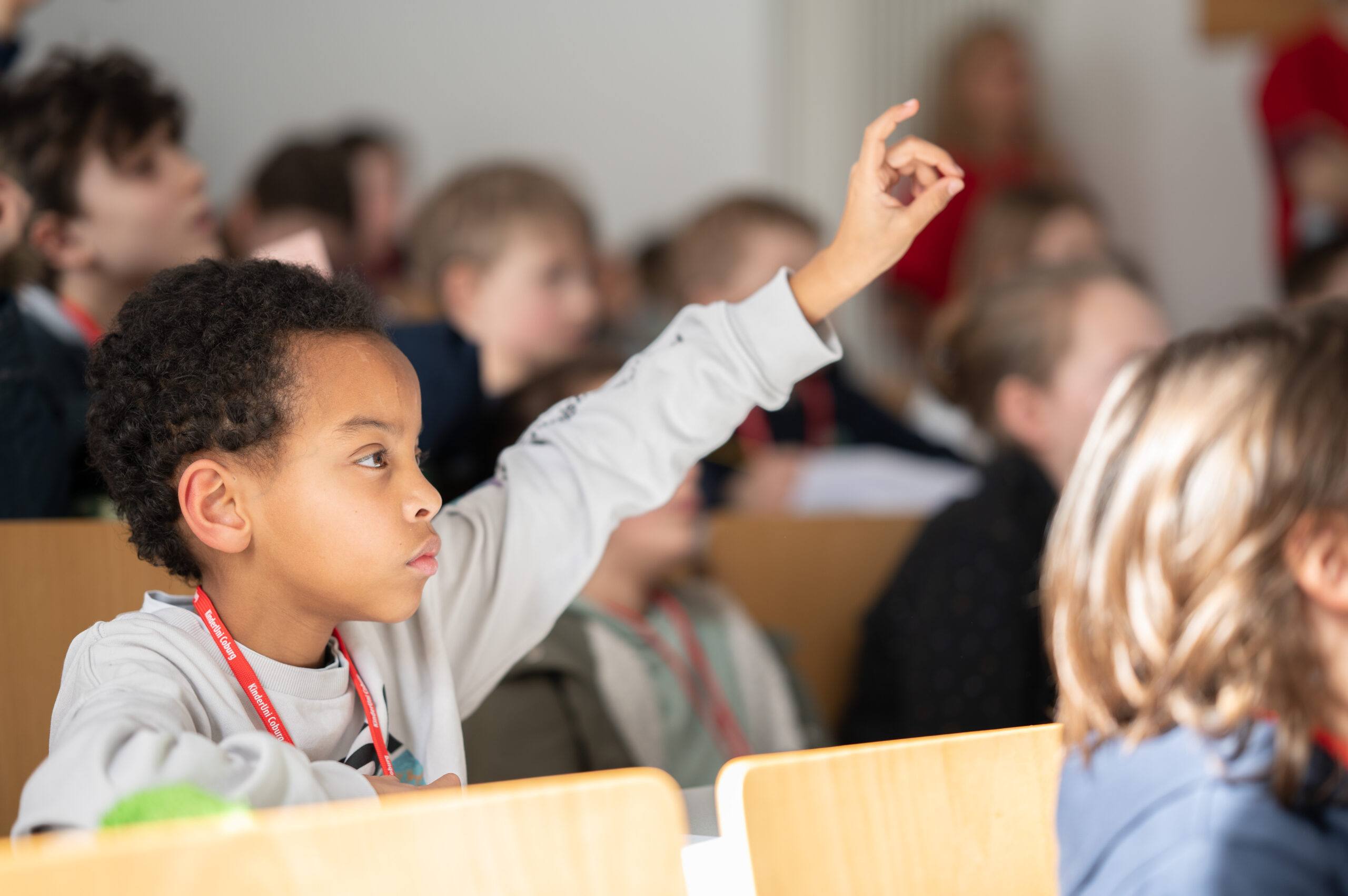 In einem hellen Hörsaal der Hochschule Coburg hebt ein junger Student in einem grauen Hemd seine Hand, sichtlich interessiert und aufmerksam. Die anderen Studenten sind ebenso konzentriert und in den Unterricht vertieft.