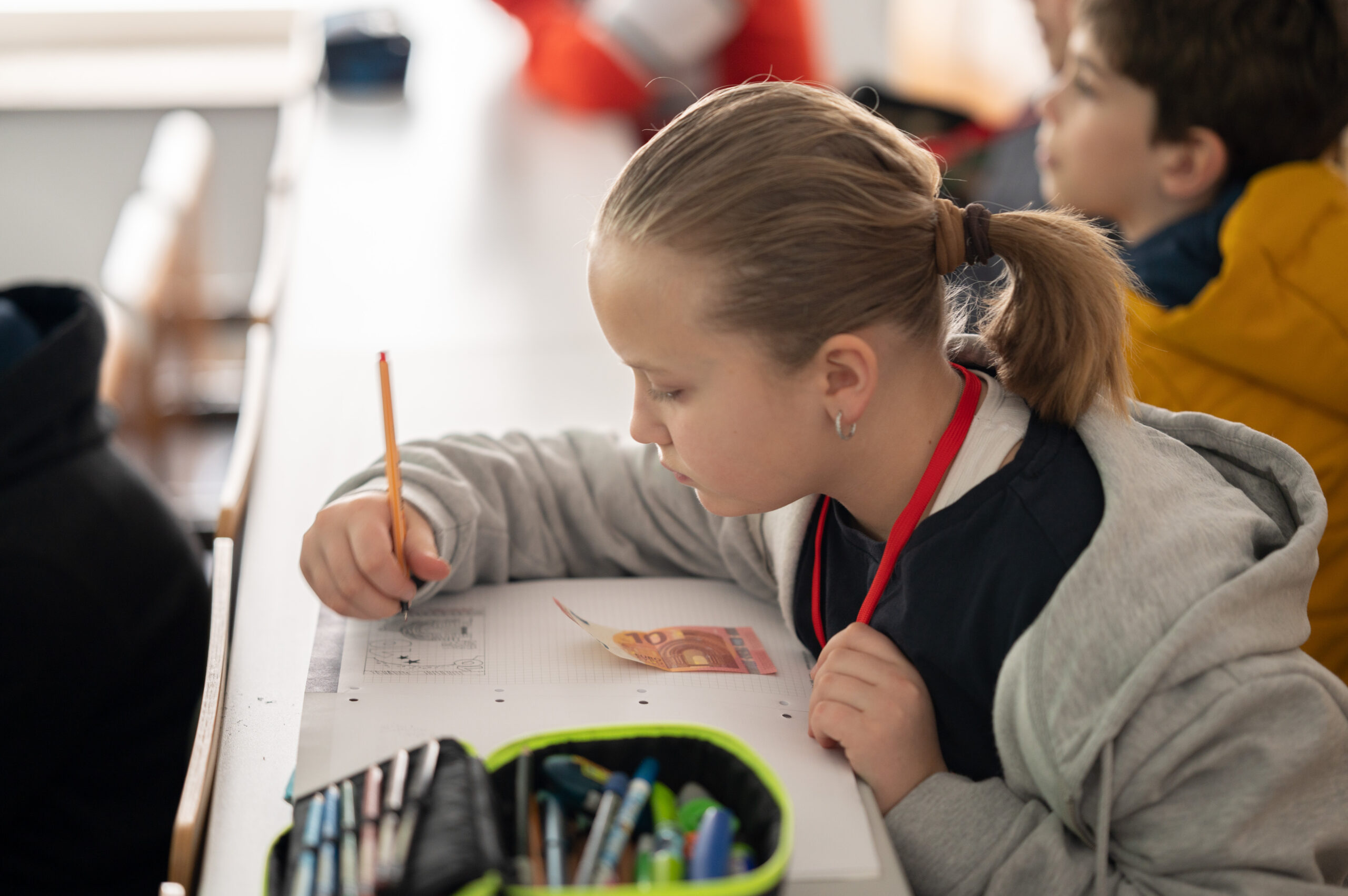 Ein junges Mädchen mit Pferdeschwanz schreibt konzentriert in ein Notizbuch an ihrem Schreibtisch an der Hochschule Coburg. Sie trägt einen grauen Kapuzenpullover und ein rotes Schlüsselband. Vor ihr liegt ein offenes Federmäppchen, während andere Studierende im Hintergrund unscharf zu sehen sind.