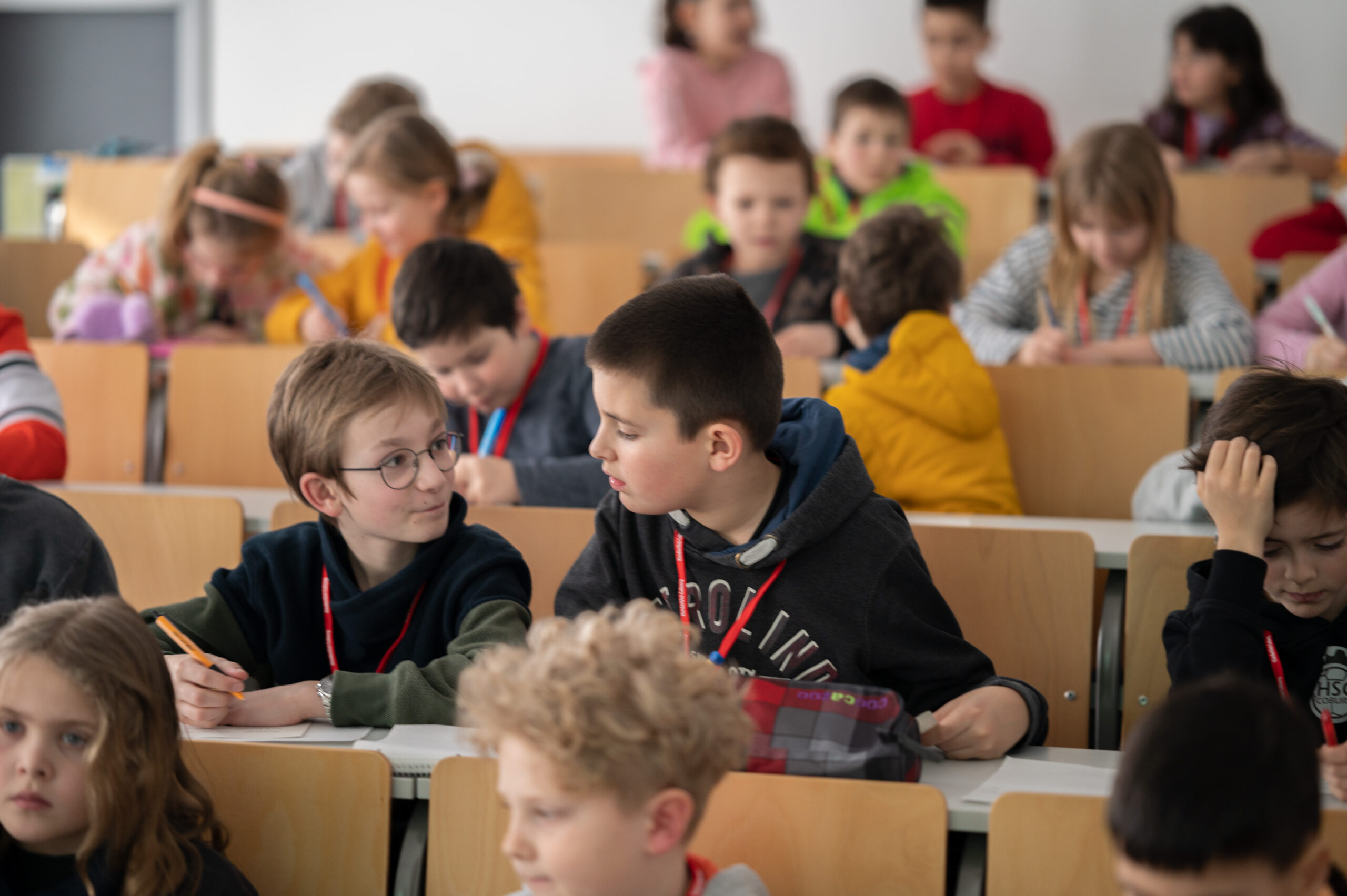 In einem lebhaften Klassenzimmer mit abgestuften Holzsitzen, die an die Hörsäle der Hochschule Coburg erinnern, herrscht bei einer Gruppe von Kindern reges Treiben. Zwei Jungen im Vordergrund unterhalten sich angeregt, einer hält einen Bleistift in der Hand, während die anderen, leger gekleidet und mit Namensschildern versehen, in ihre Aufgaben vertieft sind.