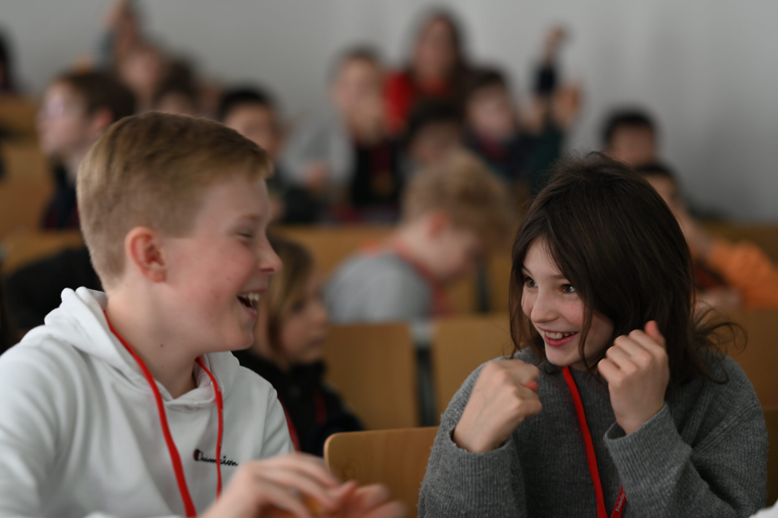 Zwei Kinder mit roten Schlüsselbändern sitzen in einem Hörsaal der Hochschule Coburg, lächeln und wirken engagiert. Im Hintergrund sitzen weitere Studierende in Reihen, etwas unscharf.