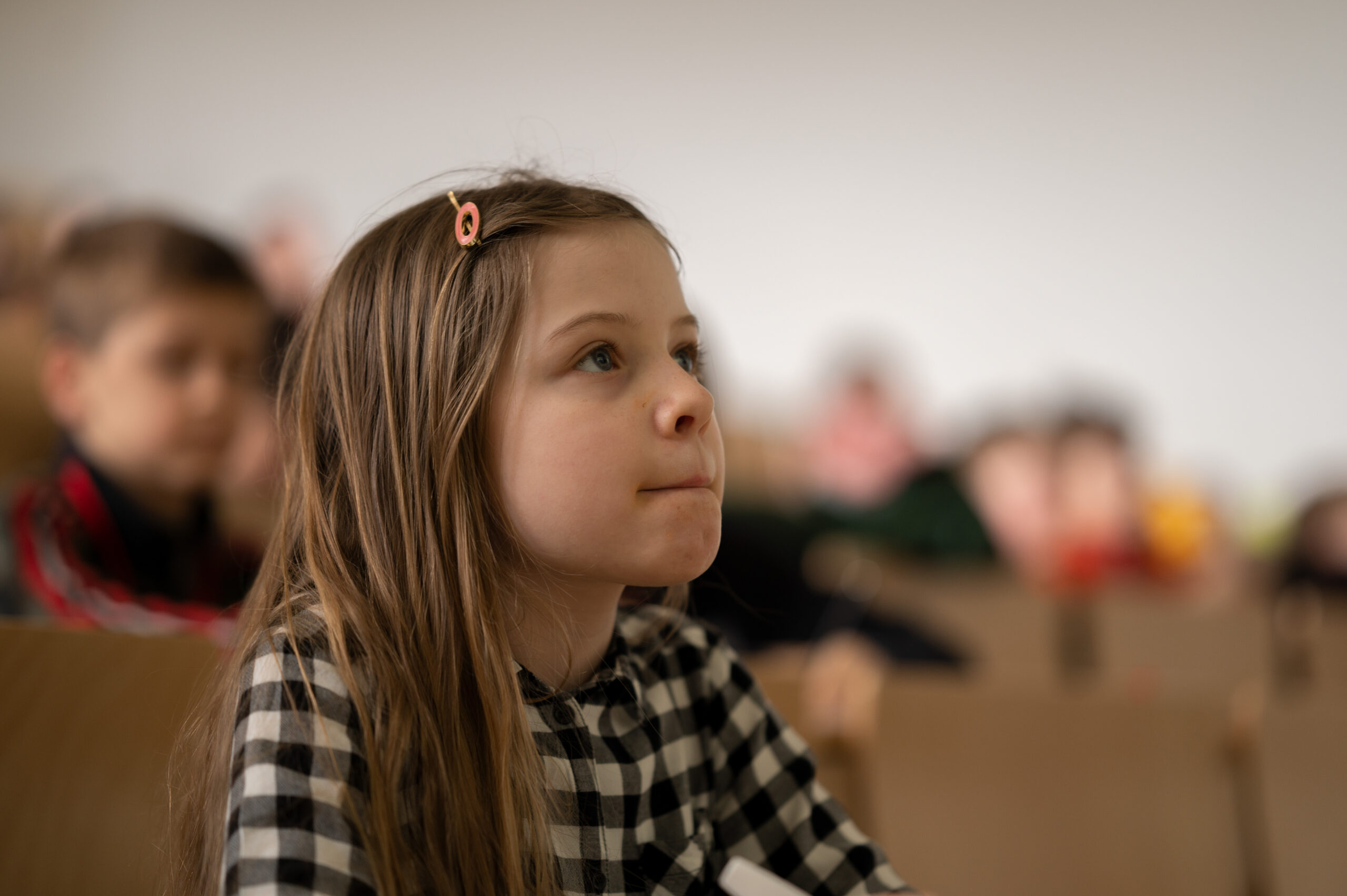 Ein junges Mädchen mit langen Haaren und einer Haarspange sitzt aufmerksam in einem Hörsaal der Hochschule Coburg. Sie trägt ein kariertes Hemd. Sie wirkt konzentriert, während im Hintergrund die anderen Studierenden unscharf zu erkennen sind, die in Reihen sitzen.