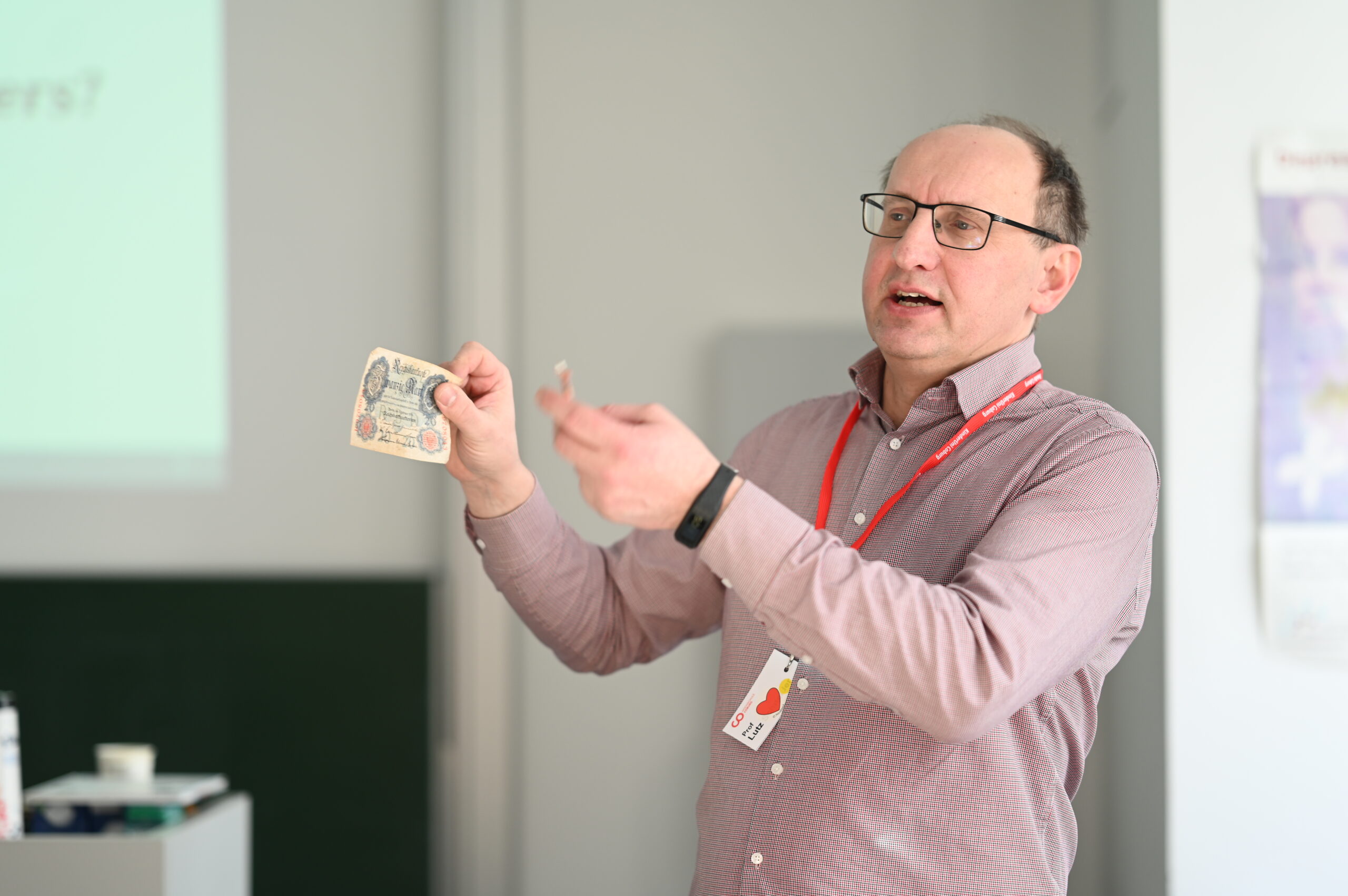 Ein Mann mit Brille und rotem Schlüsselband hält eine Banknote und einen kleinen Gegenstand hoch und bespricht diese vor einer Projektionsfläche in einem Hörsaal der Hochschule Coburg.