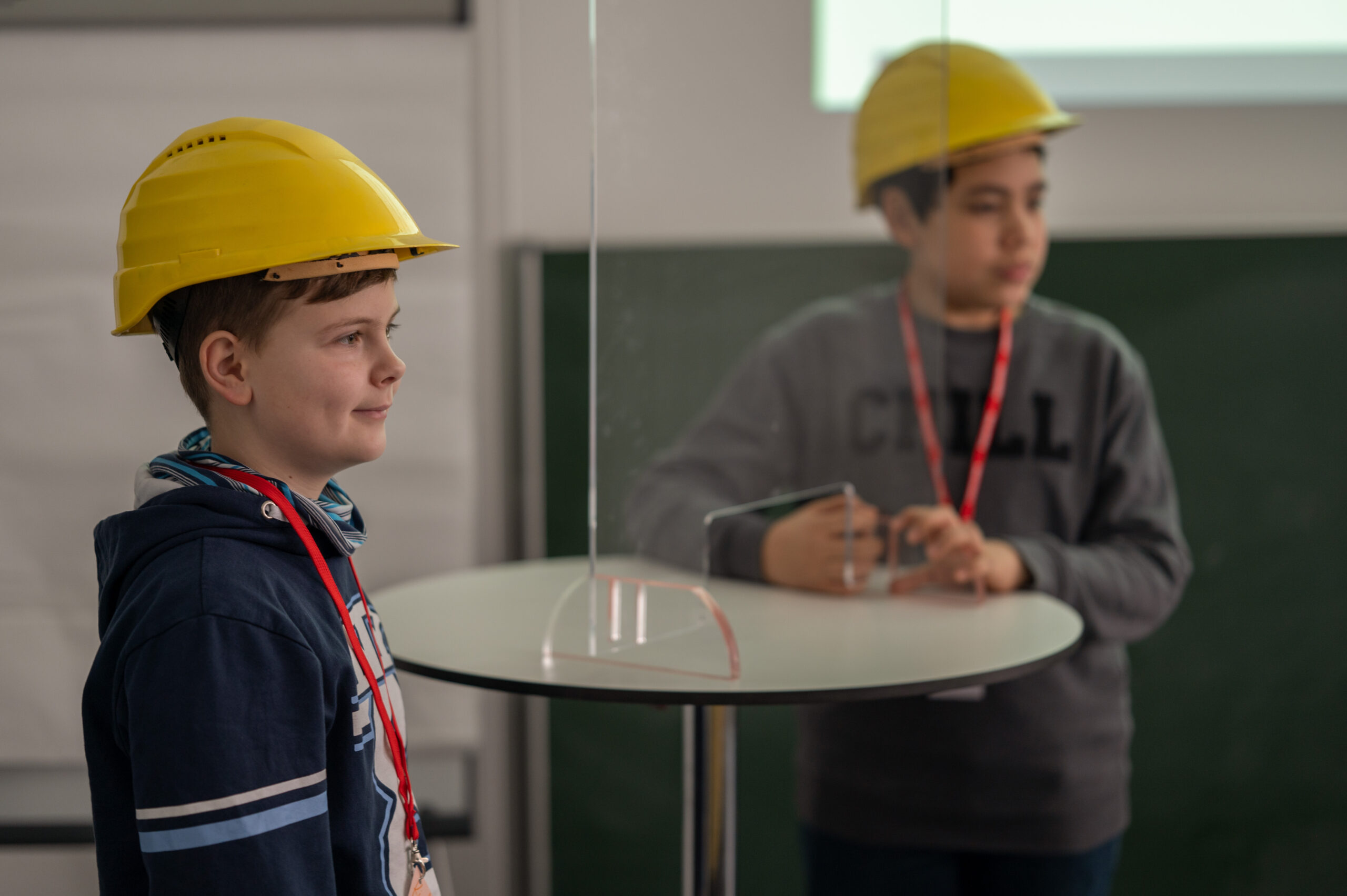 Zwei Jungen mit gelben Schutzhelmen und roten Kordeln stehen an einem Tisch mit transparenter Trennwand an der Hochschule Coburg. Einer der Jungen lächelt, während der andere ein Telefon hält. In einem Raum, in dem sowohl eine grüne Tafel als auch ein Whiteboard stehen, ist ein Raum mit einer grünen Tafel und einem Whiteboard.