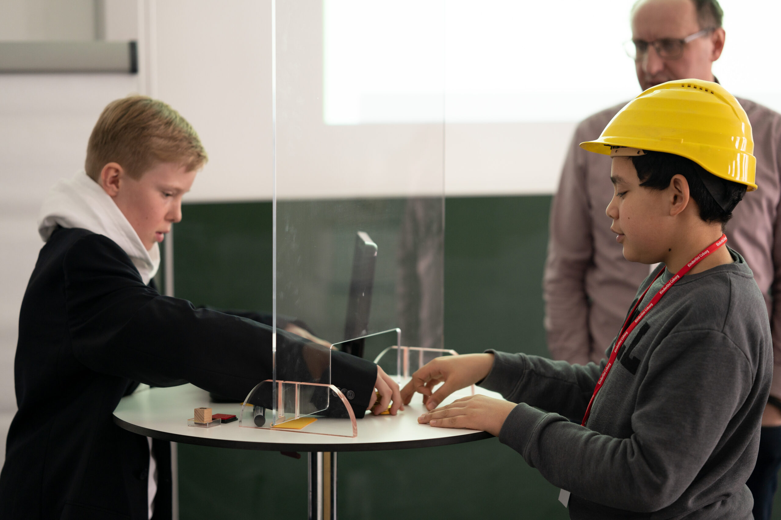 Zwei Jungen stehen an einem runden Tisch mit Glastrennwand. Einer trägt einen Anzug, der andere einen Bauhelm und Freizeitkleidung. Beide nehmen an einem wissenschaftlichen Experiment teil, das den Innovationsgeist der Hochschule Coburg widerspiegelt. Im Hintergrund beobachtet ein Erwachsener die beiden.