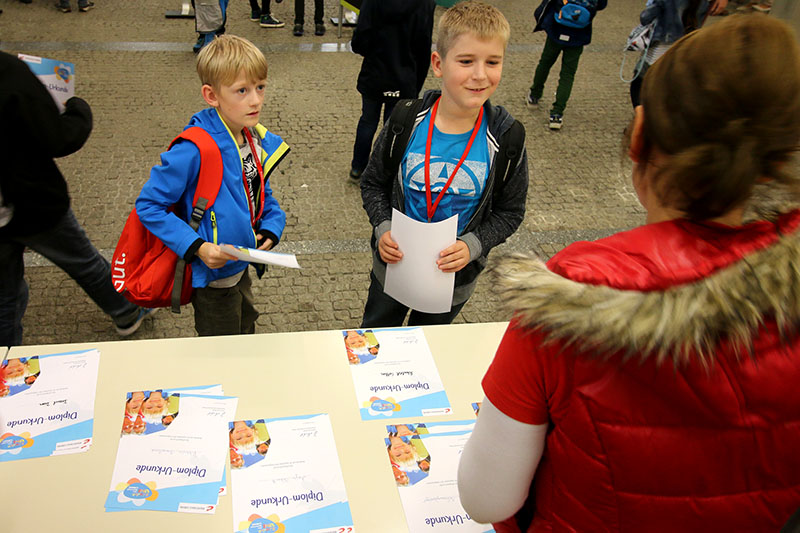 Zwei Jungen mit Lanyards stehen an einem Tisch und nehmen Papiere von einer Frau in einer roten Jacke mit Pelzbesatz entgegen. Der mit bunten Heften gefüllte Tisch scheint Teil einer Veranstaltung der Hochschule Coburg zu sein und trägt zur lebhaften Atmosphäre dieser geschäftigen Indoor-Messe bei.