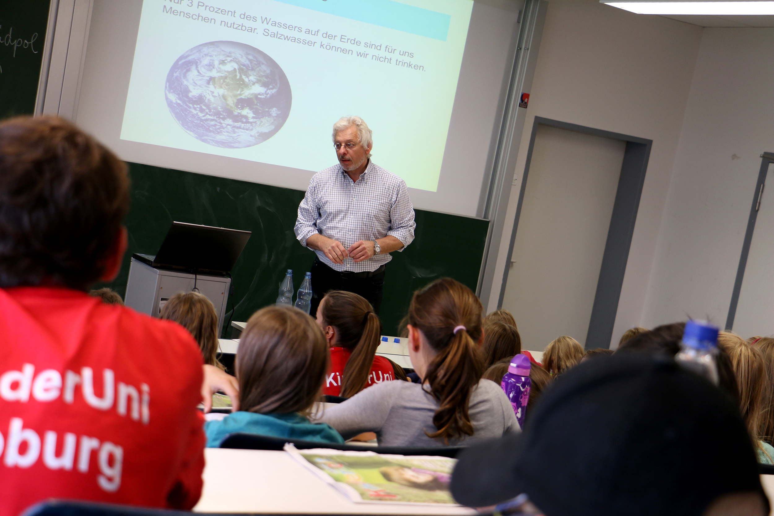 Ein Mann steht vor einem Klassenzimmer der Hochschule Coburg und spricht zu einer Gruppe von Kindern, die an ihren Tischen sitzen. Hinter ihm wird auf die Tafel ein Bild der Erde mit einem deutschen Text projiziert. Die Kinder scheinen aufmerksam und interessiert zu sein.