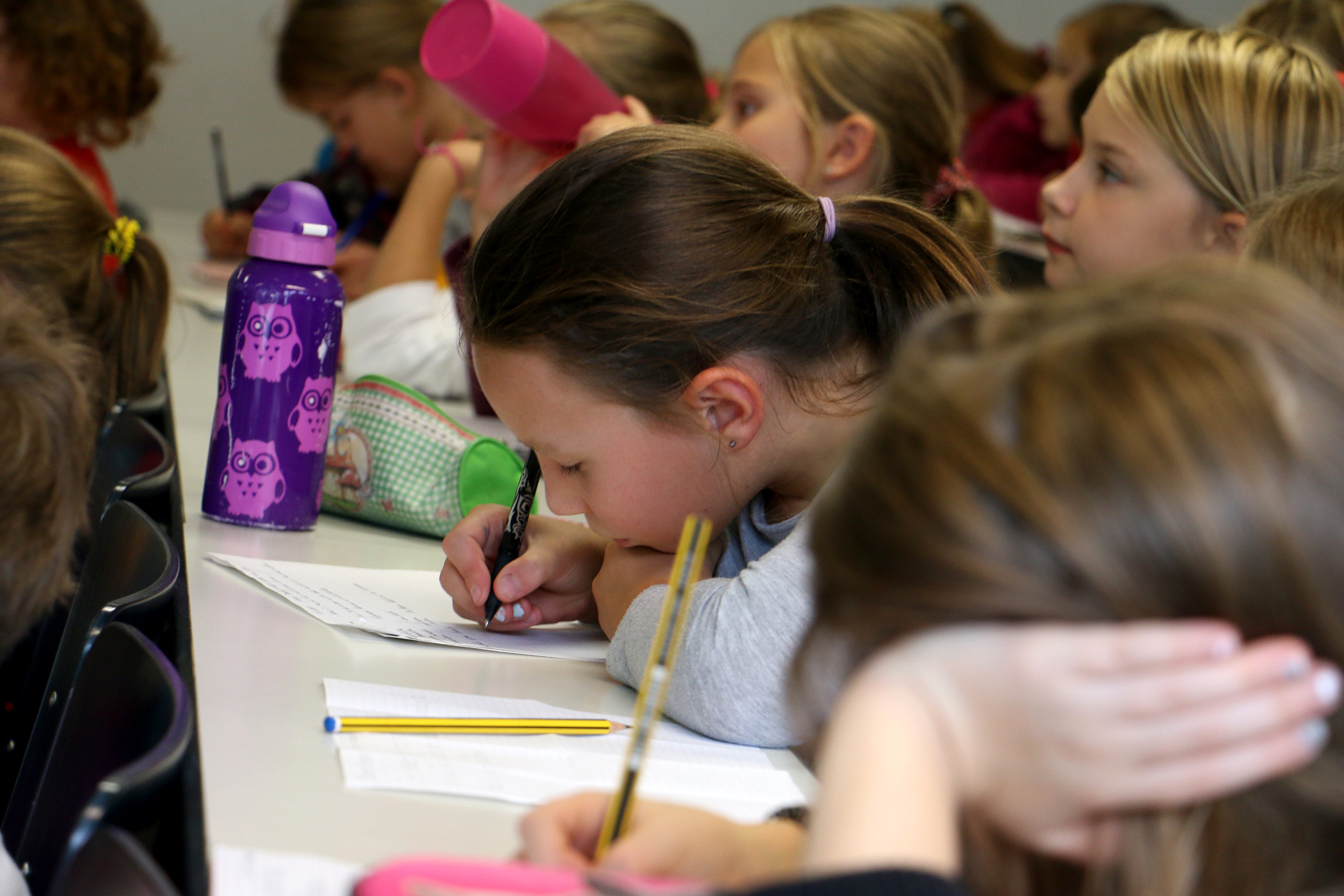 Eine Gruppe von Kindern sitzt an einem Tisch und schreibt konzentriert in Hefte an der Hochschule Coburg. Ein Mädchen im Vordergrund hält einen Bleistift und schreibt konzentriert. Bunte Wasserflaschen und Federmäppchen sind über den Tisch verteilt.