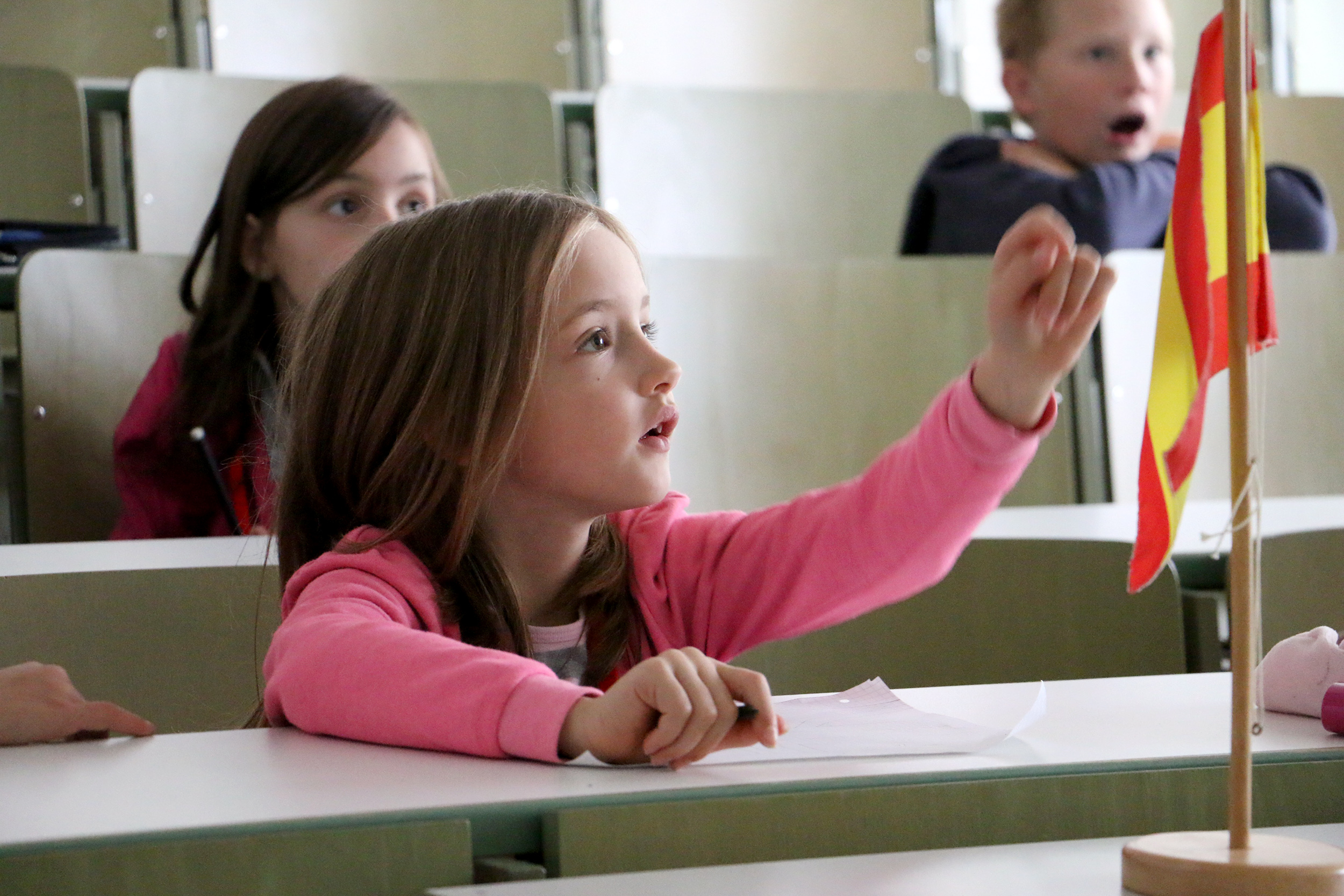 Ein junges Mädchen im rosa Shirt hebt im Hörsaal der Hochschule Coburg aufmerksam und interessiert die Hand. Sie sitzt neben einer kleinen Flagge auf dem Tisch. Im Hintergrund sitzen auch andere Kinder, eines sieht überrascht aus.