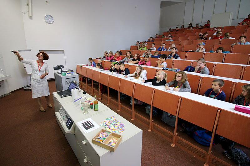 Eine Frau unterrichtet in einem großen Hörsaal der Hochschule Coburg, in dem viele Studierende an ihren Tischen sitzen. Die Studierenden hören aufmerksam zu, und auf dem Tisch ganz vorne sind verschiedene Unterrichtsmaterialien zu sehen.