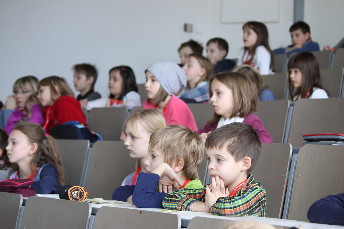 Eine Gruppe kleiner Kinder sitzt in einem Hörsaal der Hochschule Coburg und verfolgt aufmerksam eine Präsentation. Sie sitzen in mehreren Reihen, manche stützen ihr Kinn auf die Hände. Der Raum ist mit Tageslicht gut beleuchtet.