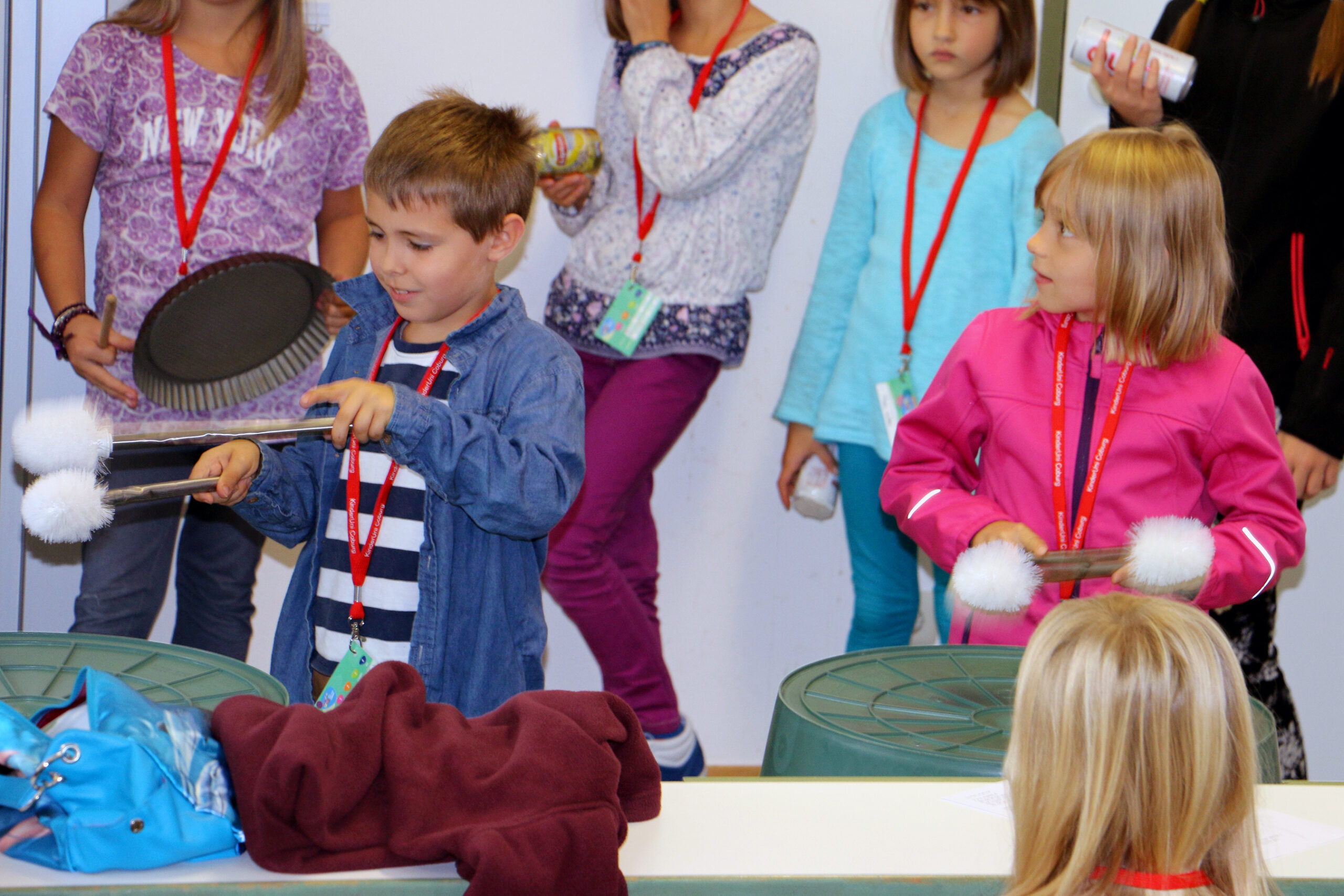 Kinder der Hochschule Coburg beteiligen sich an einer lebhaften Musikaktivität, bei der sie Küchenutensilien als Trommeln verwenden. Zwei Kinder mit Schlägeln stehen davor und spielen auf großen Behältern, während andere in Freizeitkleidung und mit roten Schlüsselbändern zuschauen und mitmachen. Die Szene ist lebendig und interaktiv.