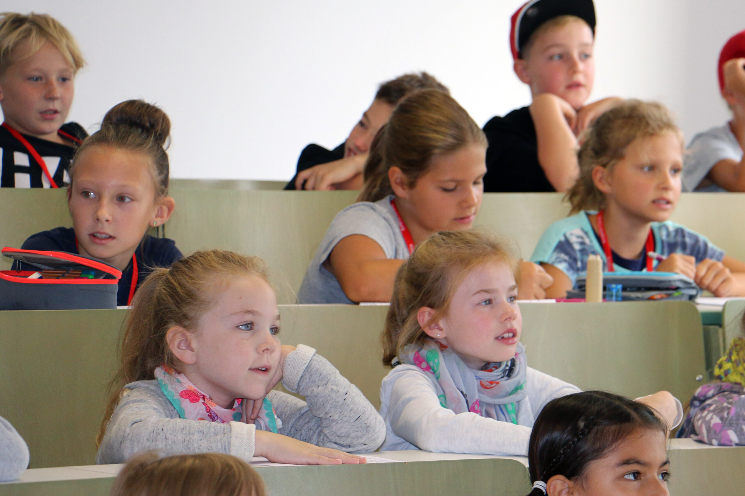 Kinder sitzen in einem Klassenzimmer und hören aufmerksam zu. Sie sitzen in abgestuften Reihen, tragen Freizeitkleidung und einige tragen bunte Schlüsselbänder. Die helle Umgebung könnte man leicht mit einem Seminar der Hochschule Coburg verwechseln, so konzentriert und energiegeladen ist der schlichte Hintergrund.