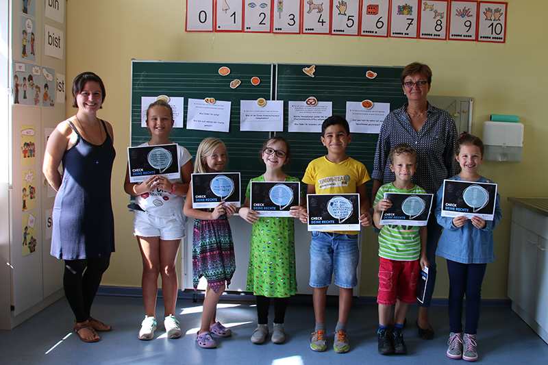 Eine Gruppe von sieben Kindern steht lächelnd in einem Klassenzimmer der Hochschule Coburg. Jedes Kind hält ein Zertifikat in der Hand. Zwei Erwachsene stehen neben ihnen und strahlen vor Stolz. Hinter ihnen ist eine Tafel mit Zeichnungen und Texten. Der Raum ist gut beleuchtet und mit pädagogischen Plakaten dekoriert.