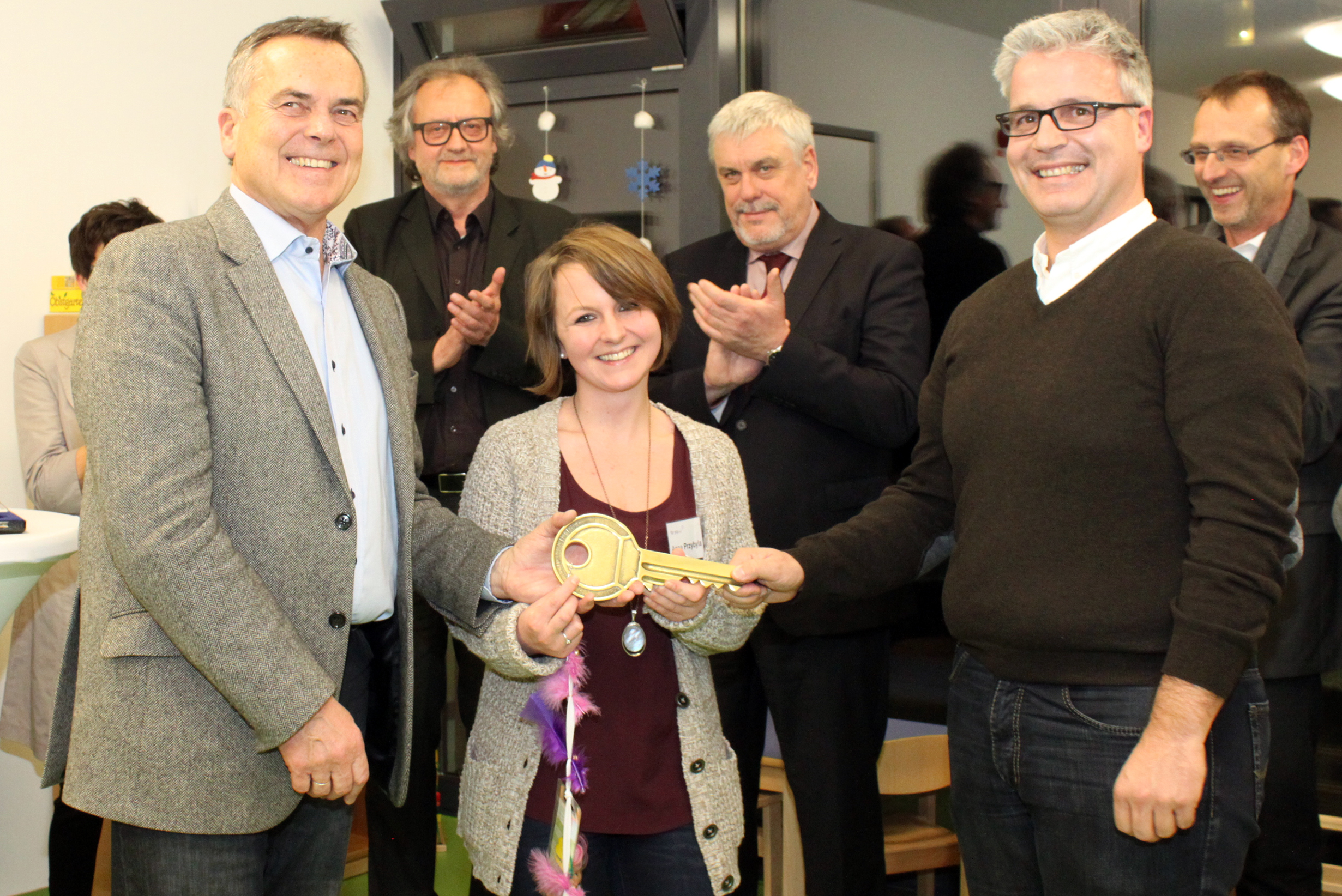 Eine Gruppe von Menschen lächelt, während eine Frau einen großen Zeremonienschlüssel hält, neben ihr stehen zwei Männer. Andere im Hintergrund klatschen und schauen zu, was auf eine feierliche Veranstaltung in geschlossenen Räumen schließen lässt, möglicherweise an der Hochschule Coburg.