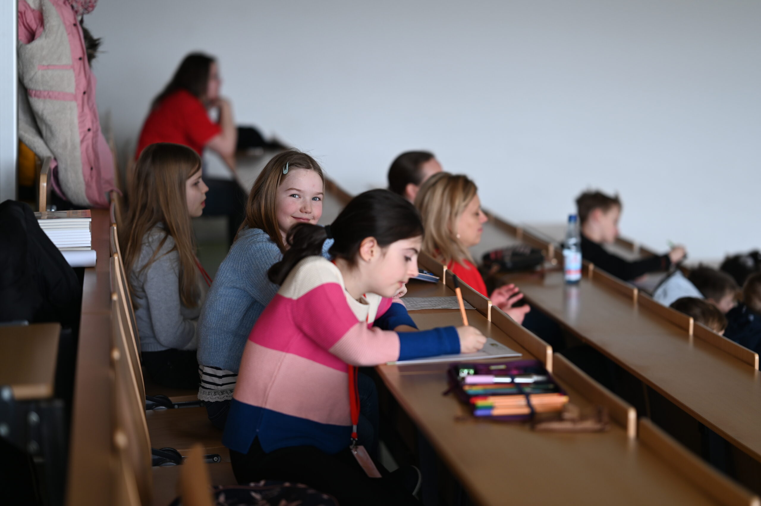 In einem Hörsaal der Hochschule Coburg sitzen Studierende an ihren Schreibtischen. Ein junges Mädchen in einem rosa Pullover schreibt, während die anderen um sie herum reden oder aufmerksam bleiben. Die Atmosphäre wirkt locker, aber dennoch konzentriert auf die anstehende Aufgabe.