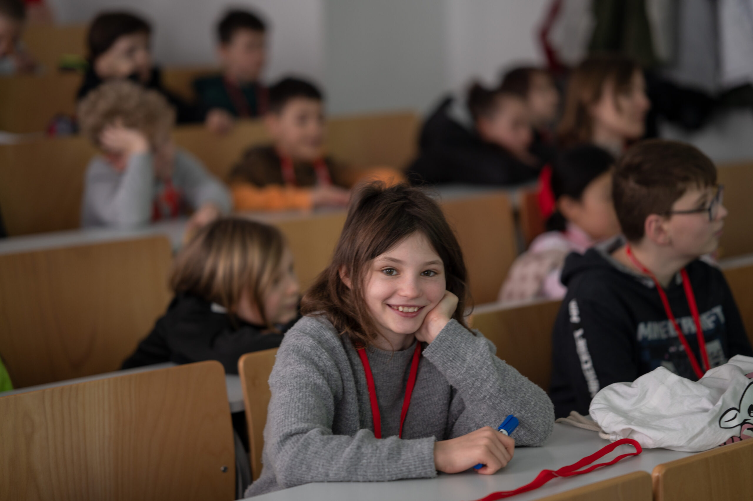 Kinder sitzen in einem Hörsaal der Hochschule Coburg und hören einer Vorlesung zu. Ein Mädchen im Vordergrund lächelt und hält einen Stift in der Hand. Es trägt einen grauen Pullover mit einem roten Schlüsselband. Andere Studenten sitzen in Reihen hinter ihr und hören aufmerksam zu. Der Raum ist mit Holzstühlen und Tischen ausgestattet.