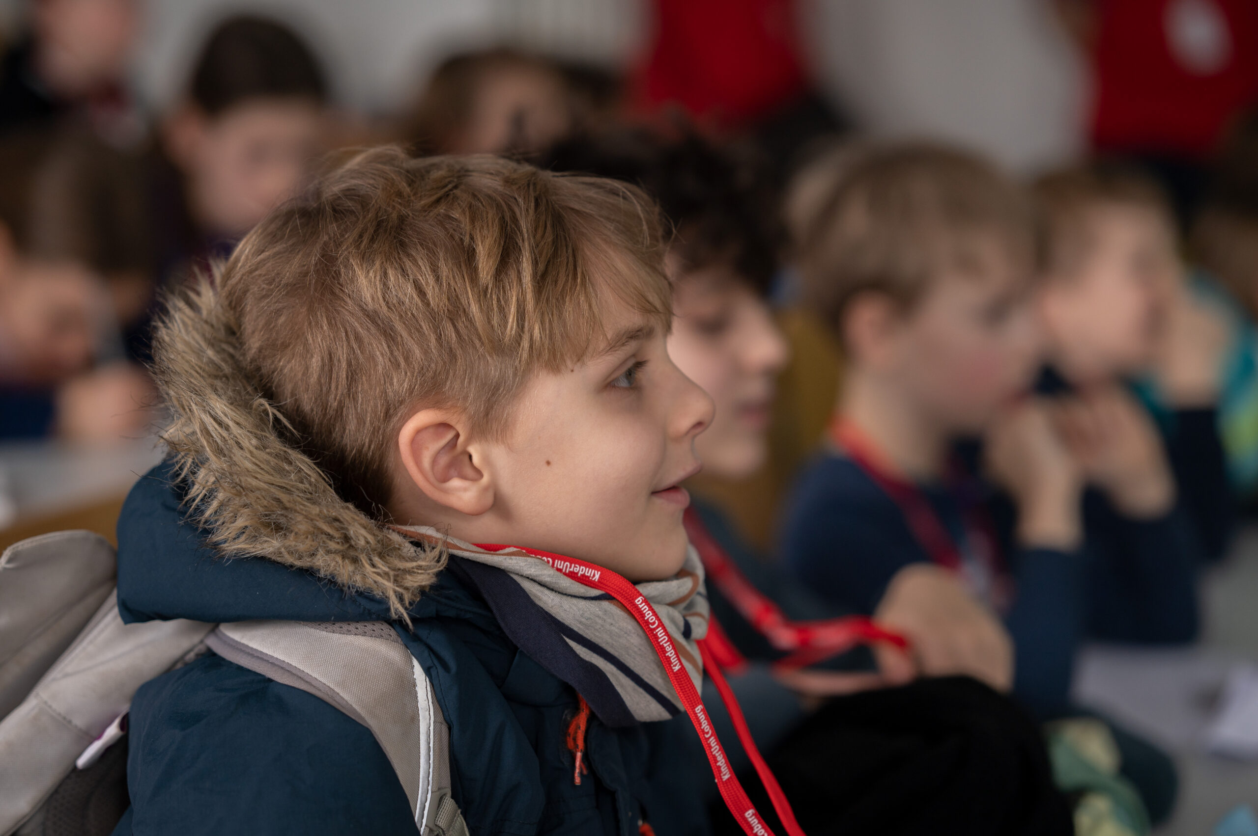 Ein kleiner Junge in einem blauen Mantel mit pelzbesetzter Kapuze sitzt aufmerksam in einem Hörsaal der Hochschule Coburg. Um den Hals trägt er ein rotes Band und ist von anderen Schülern umgeben, die ebenfalls konzentriert dem Unterricht zuhören.