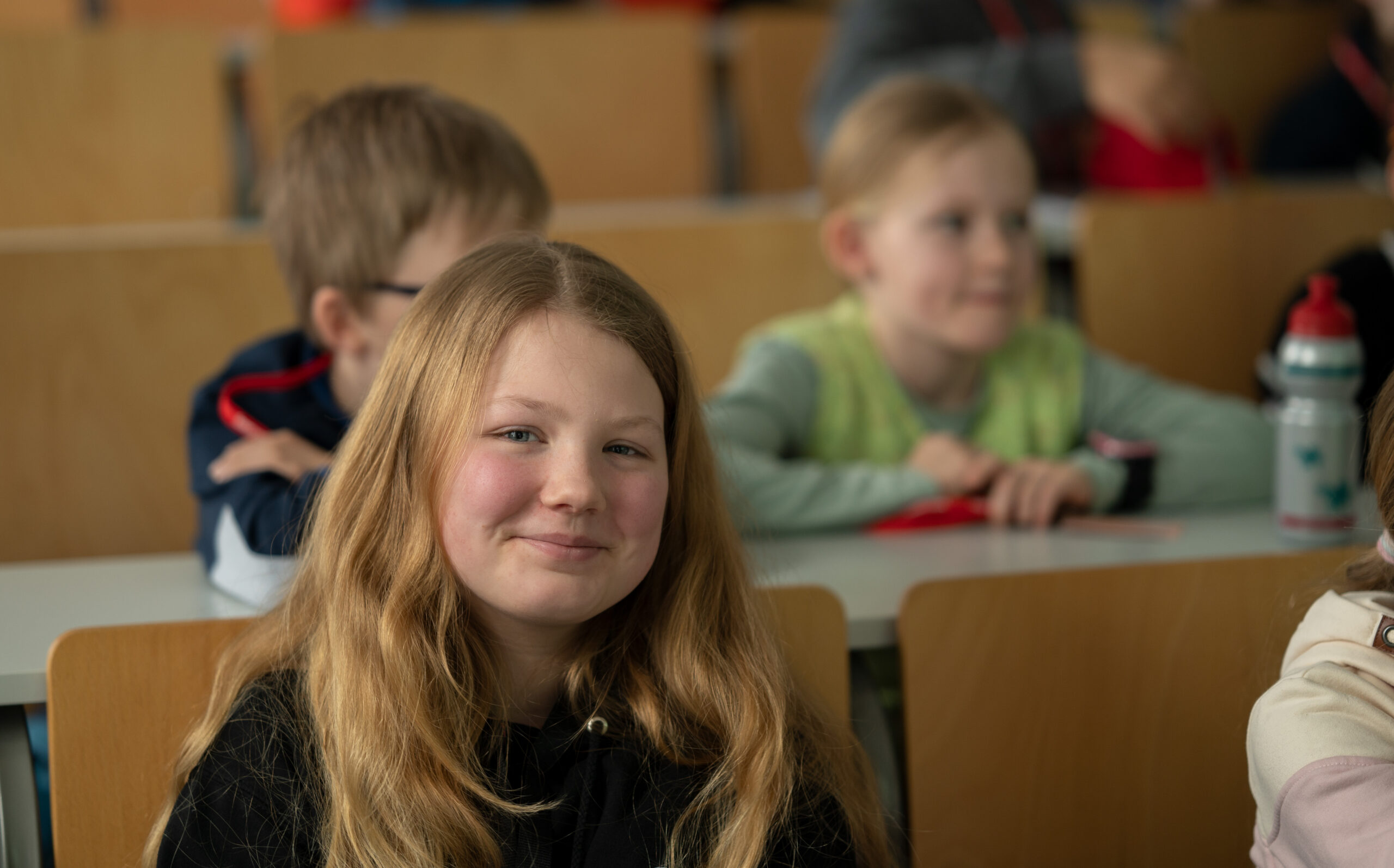 Ein junges Mädchen mit langen blonden Haaren sitzt lächelnd in einem Klassenraum der Hochschule Coburg. Hinter ihr sitzen weitere Kinder an ihren Tischen und scheinen sich auf etwas zu konzentrieren. Der Klassenraum ist mit Holzstühlen ausgestattet und hat eine helle Atmosphäre.