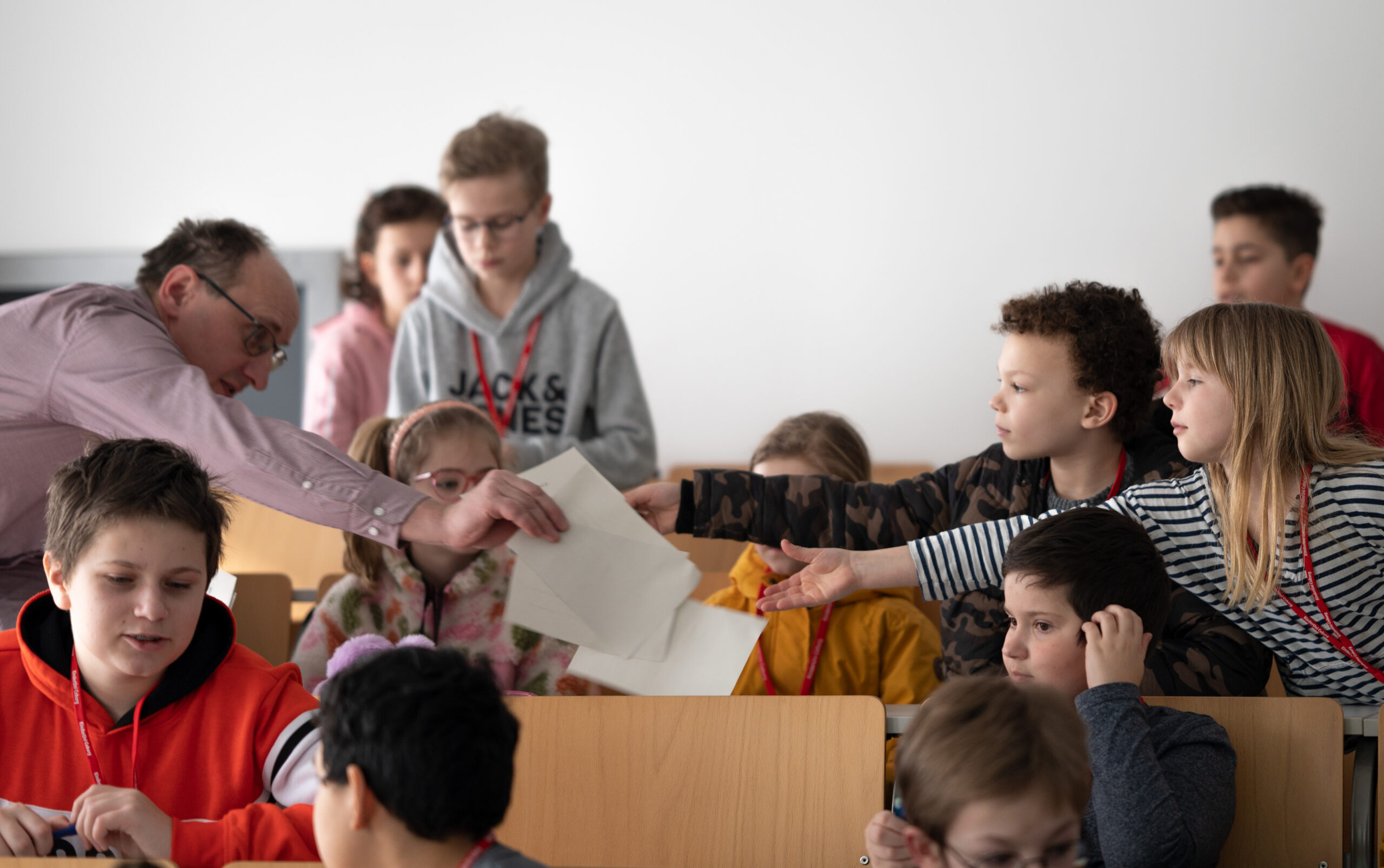 Ein Lehrer der Hochschule Coburg verteilt Blätter an eine gemischte Gruppe von Schülern, die in einem Klassenraum sitzen. Die Kinder, die leger gekleidet sind, konzentrieren sich auf die Aktivität und interagieren in dem gut beleuchteten Raum mit neutralem Hintergrund miteinander.