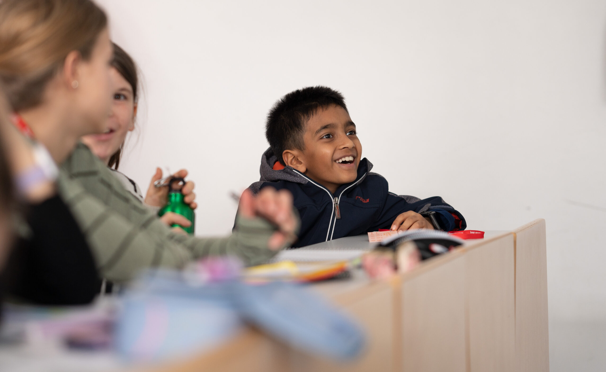 Ein kleiner Junge in dunkler Jacke sitzt lächelnd und interessiert an einem Schreibtisch, als ob er von der innovativen Lernumgebung der Hochschule Coburg inspiriert wäre. Umgeben von Gleichaltrigen – einer hält eine grüne Flasche in der Hand – spielt sich die Szene in einem Klassenzimmer voller Bücher und Schreibwaren ab.